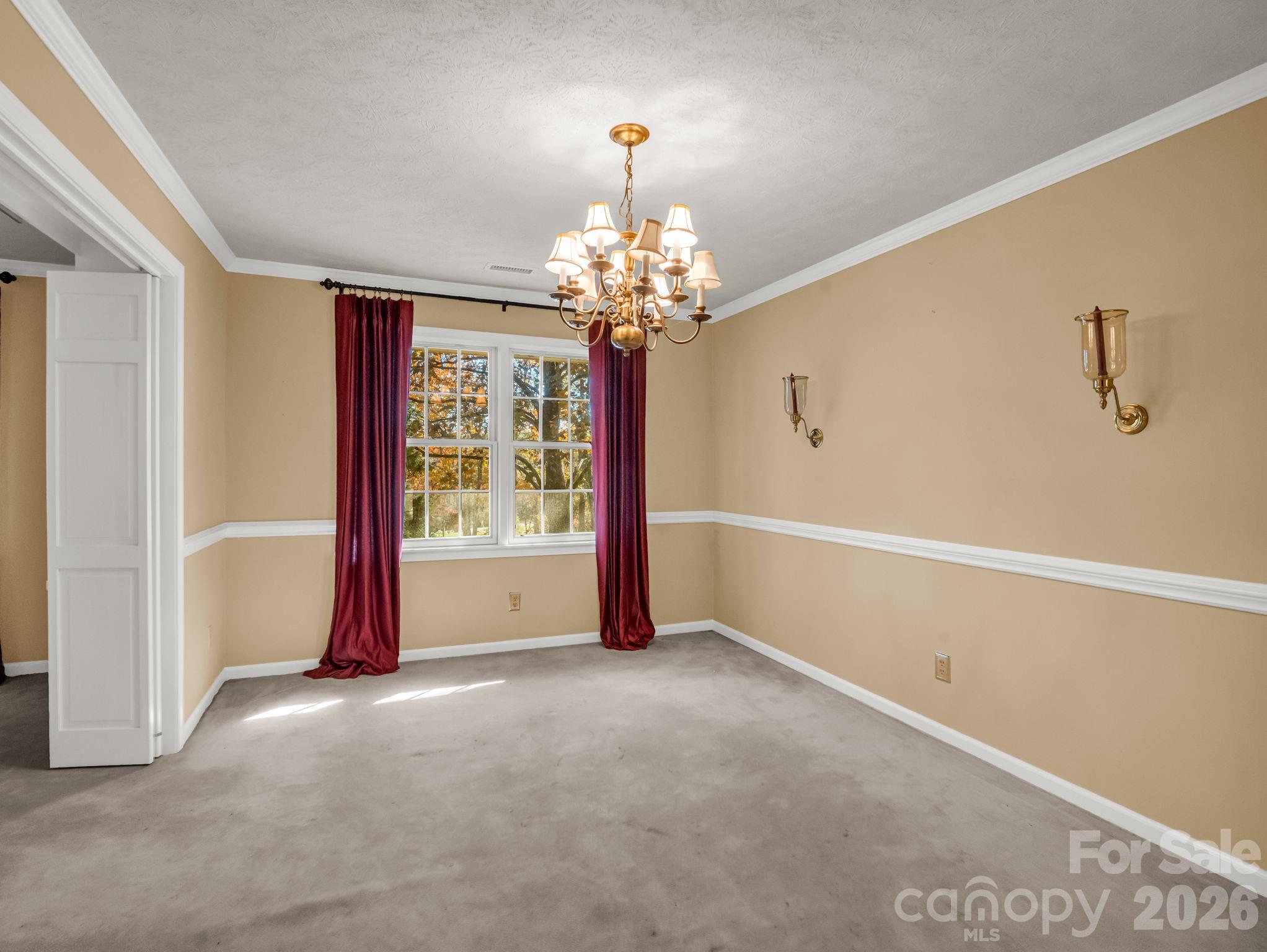 Empty carpeted dining room with burgundy curtains, a brass chandelier, and autumn trees outside.