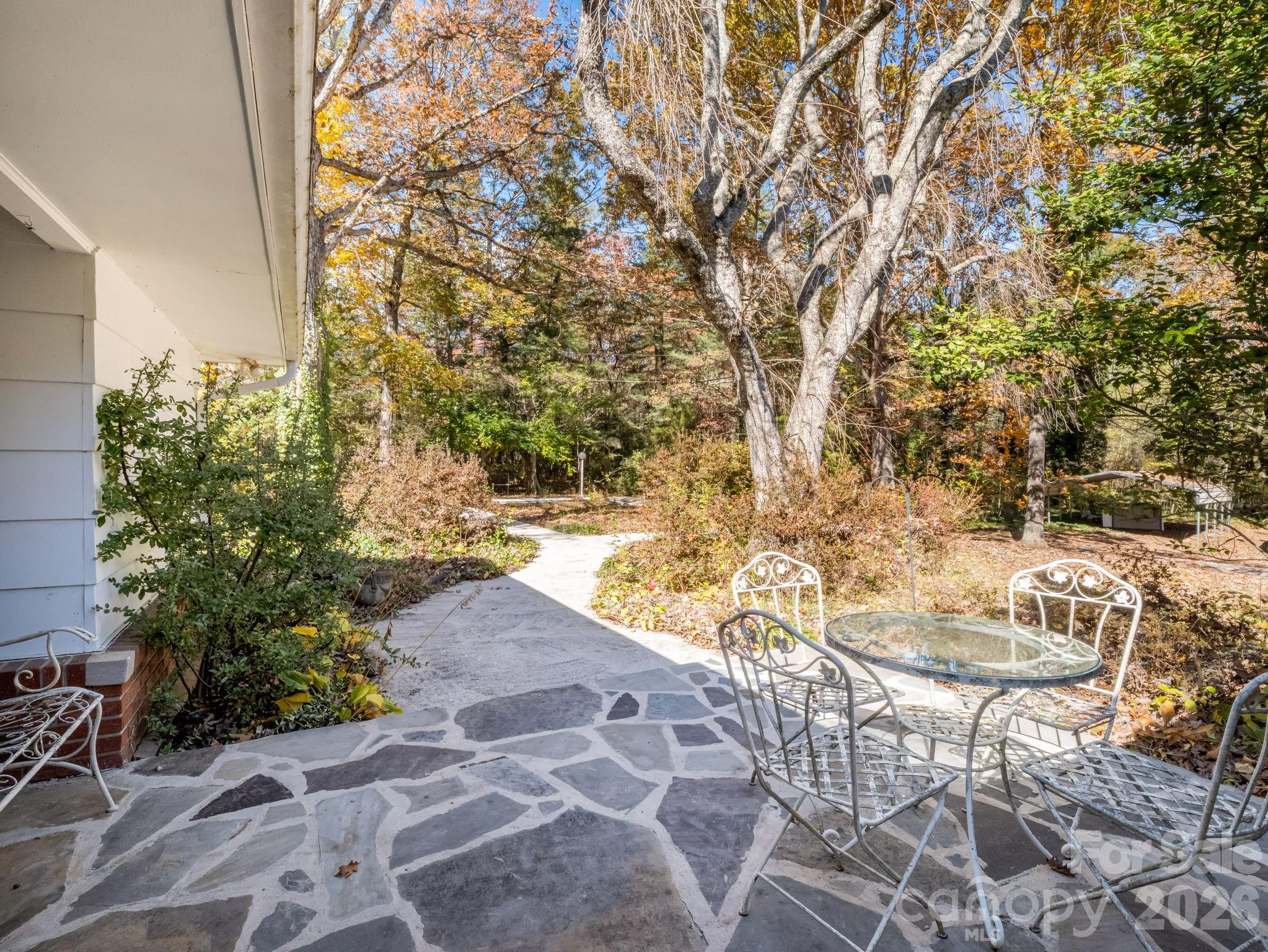Stone patio with a glass-top table and metal chairs beside a wooded garden with autumn foliage.