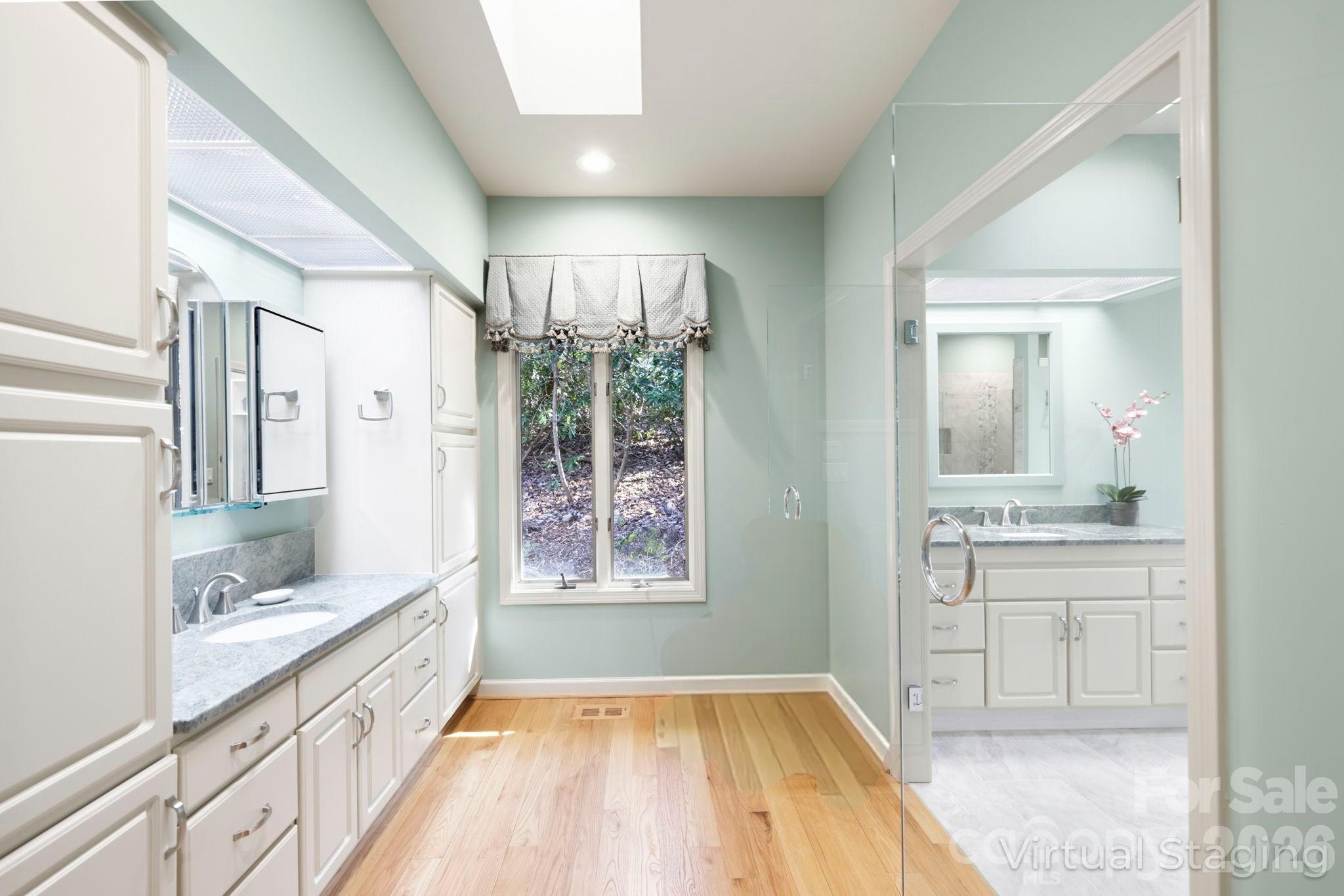 Bathroom with white double-sink vanity, glass shower, hardwood flooring, and a window overlooking trees.