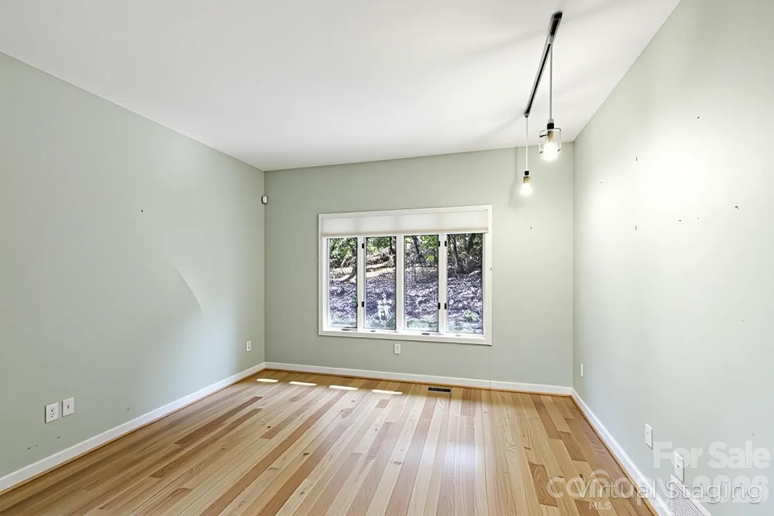 Empty room with hardwood floors, pale green walls, pendant lights, and a wide window overlooking trees.