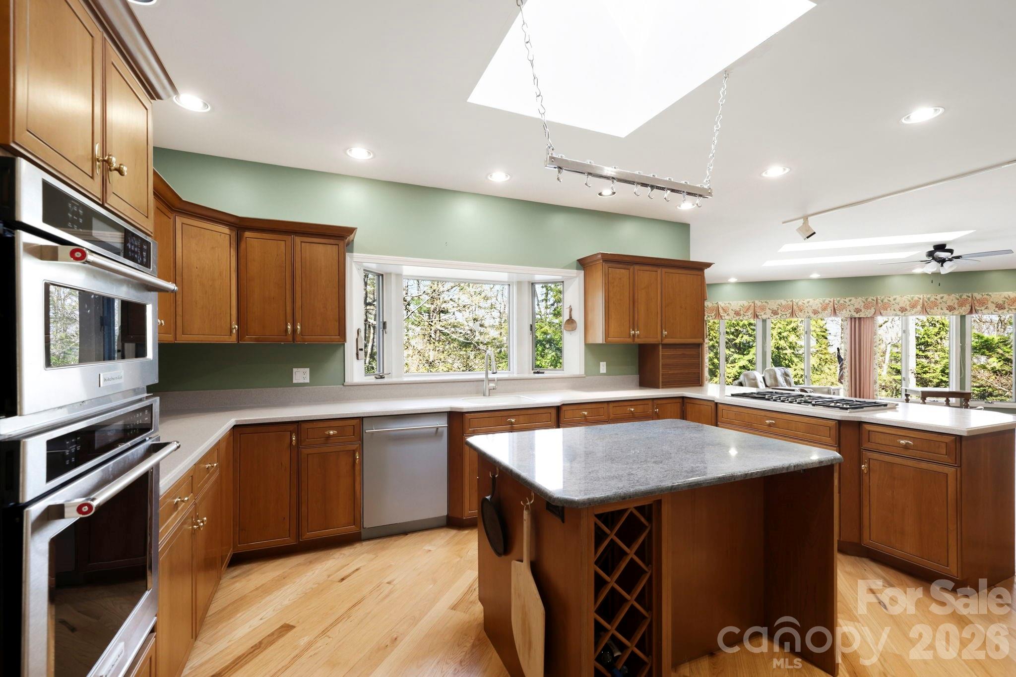 Kitchen with warm wood cabinets, a large stone island, stainless steel double ovens, and broad windows.