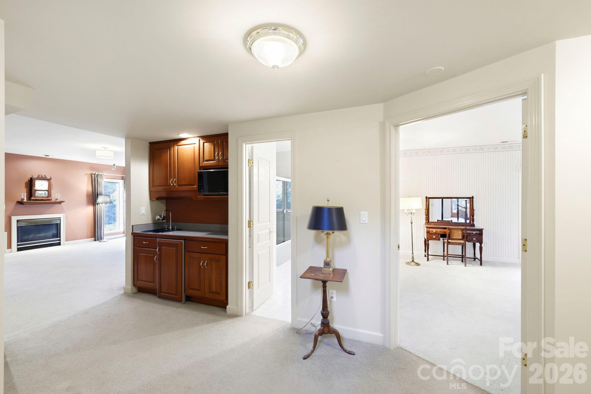 Kitchen with dark wood cabinets between a carpeted living room with fireplace and a room with a vanity.