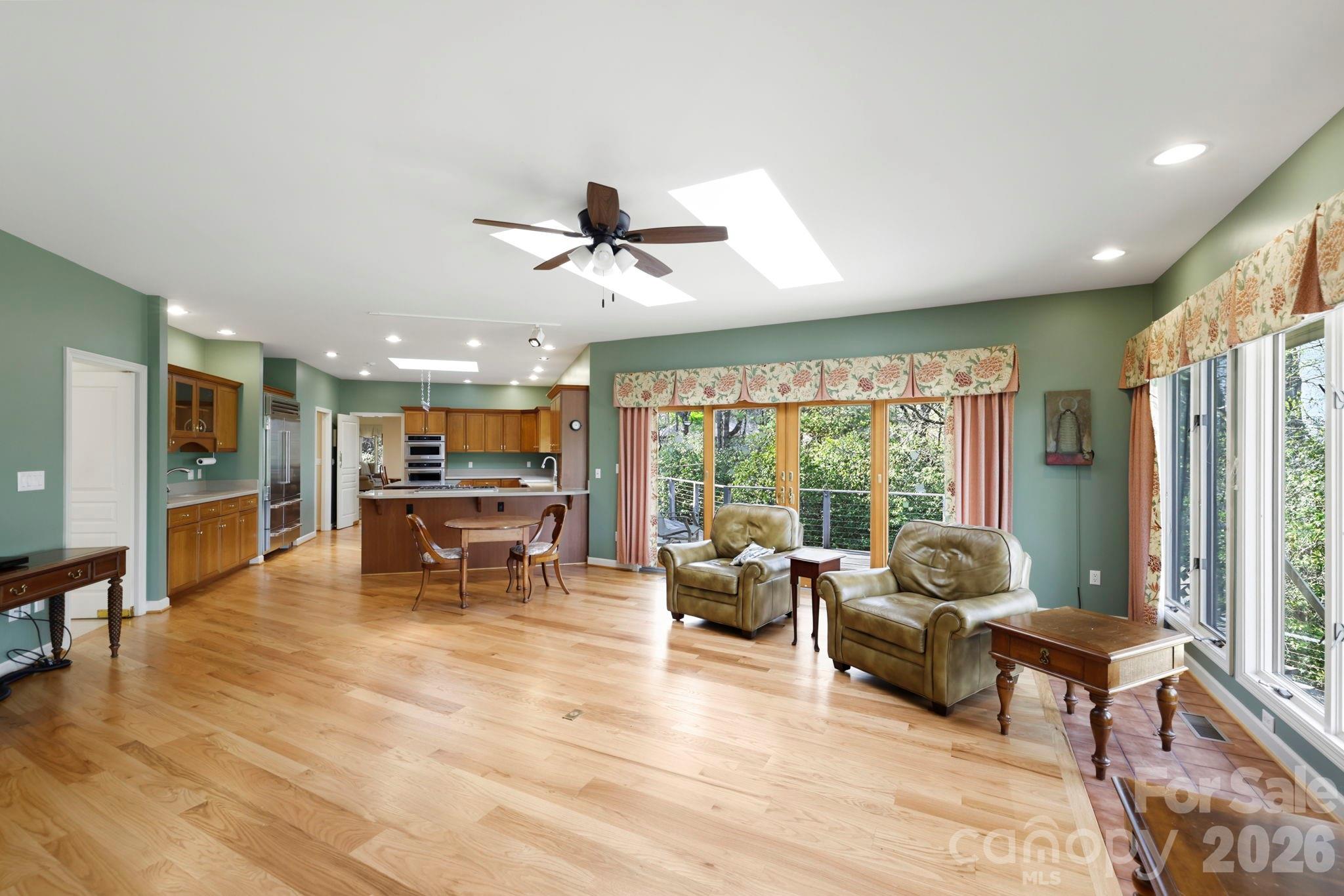 Open-plan living room and kitchen with hardwood floors, sage-green walls, and broad windows overlooking trees.