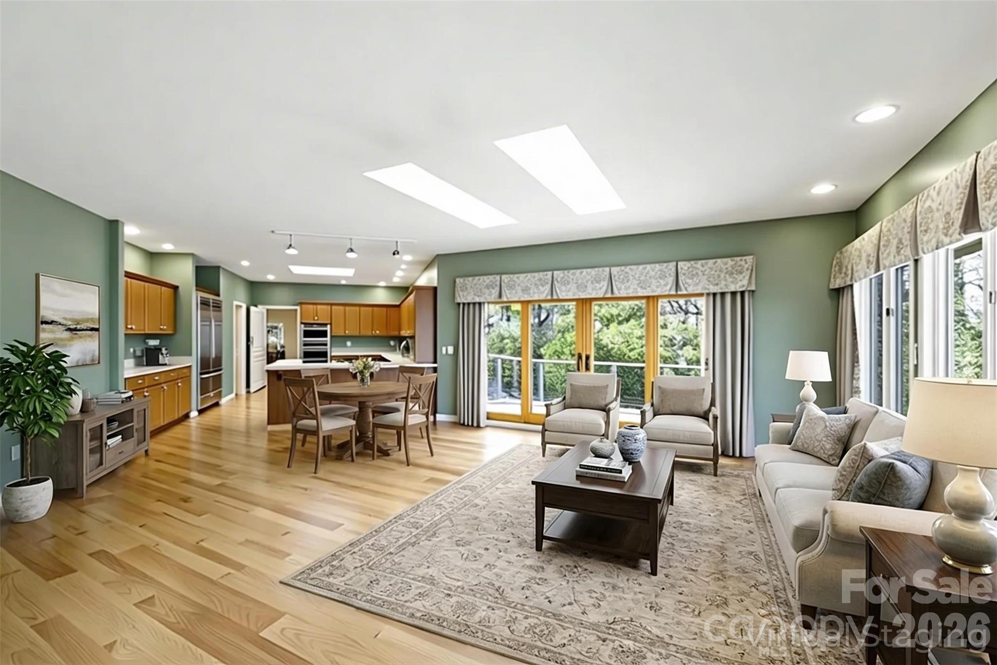 Open-plan living room and kitchen with hardwood floors, green walls, and large windows overlooking trees.