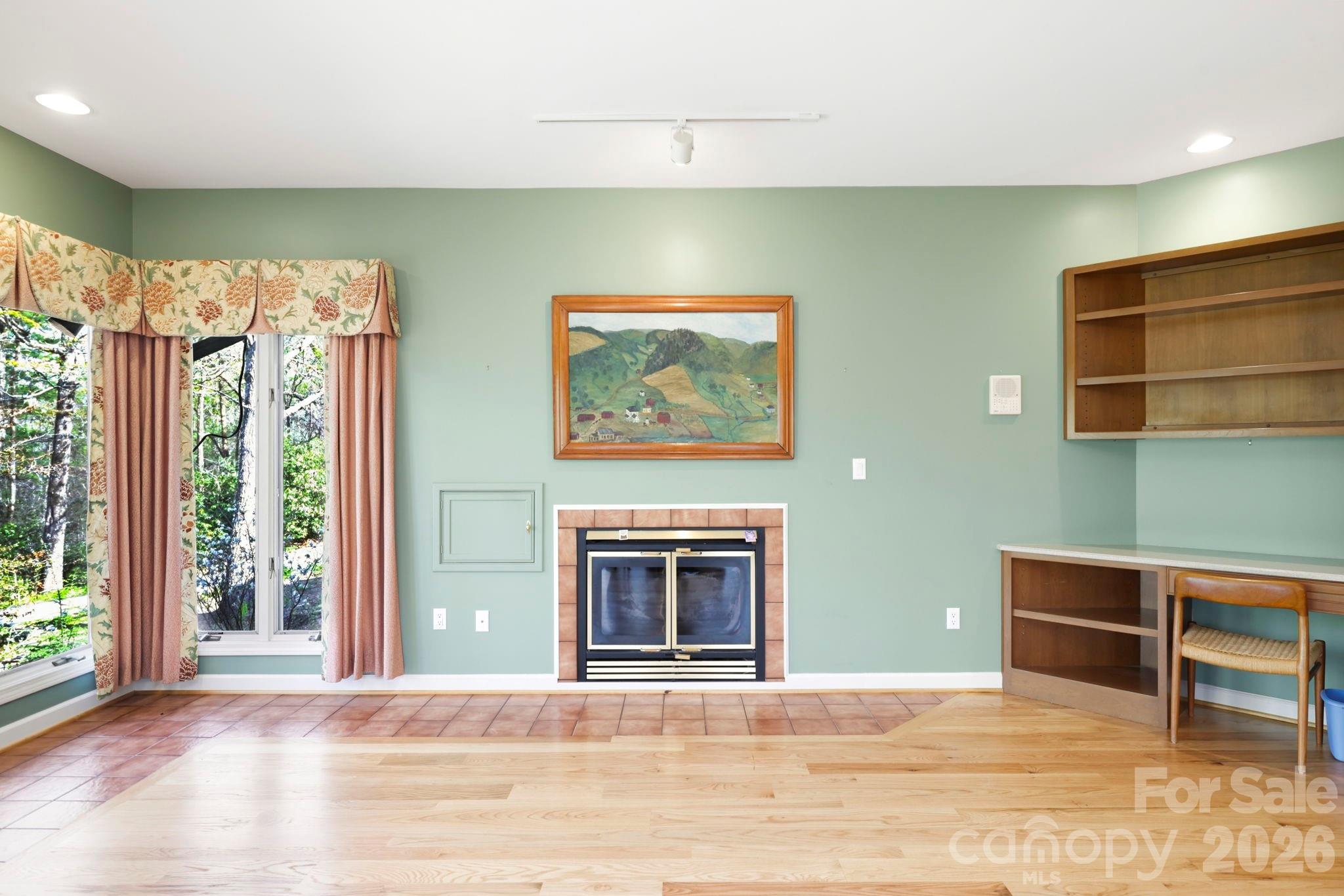 Living room with a fireplace, built-in wood shelving, and windows overlooking trees.