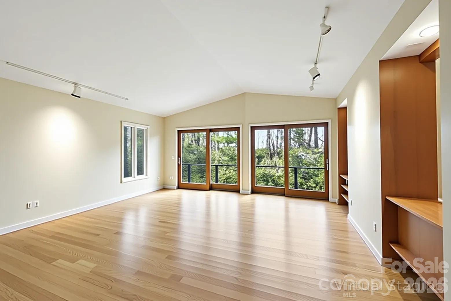 Empty room with light wood floors, track lighting, built-in shelving, and sliding glass doors overlooking trees.