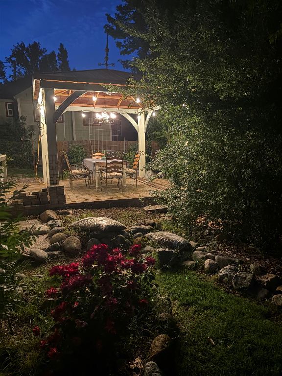 Illuminated backyard patio with a covered dining area, rock garden, and red flowers at night.