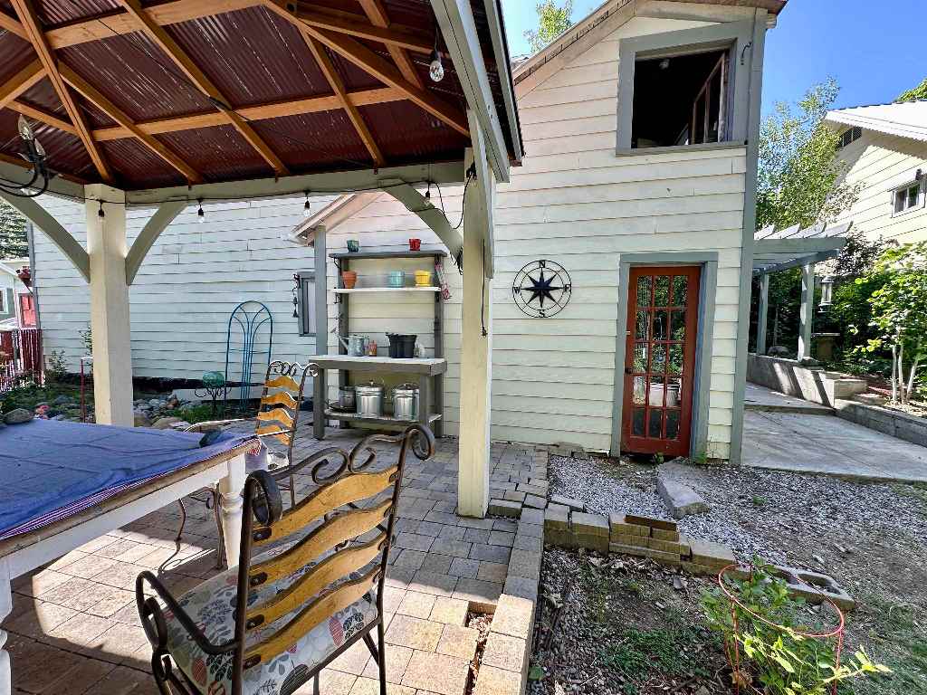 Covered patio with outdoor dining furniture, pavers, and a small garden beside a red glass-paneled door.