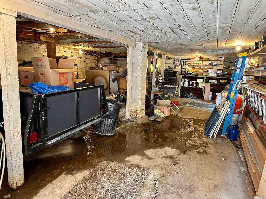 Cluttered basement garage with concrete support posts, stored tools, and puddles on the floor.