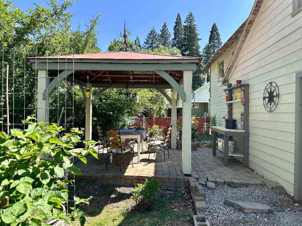 Paved patio beside a light-colored house, with a red-roofed pergola shading a dining table and chairs.