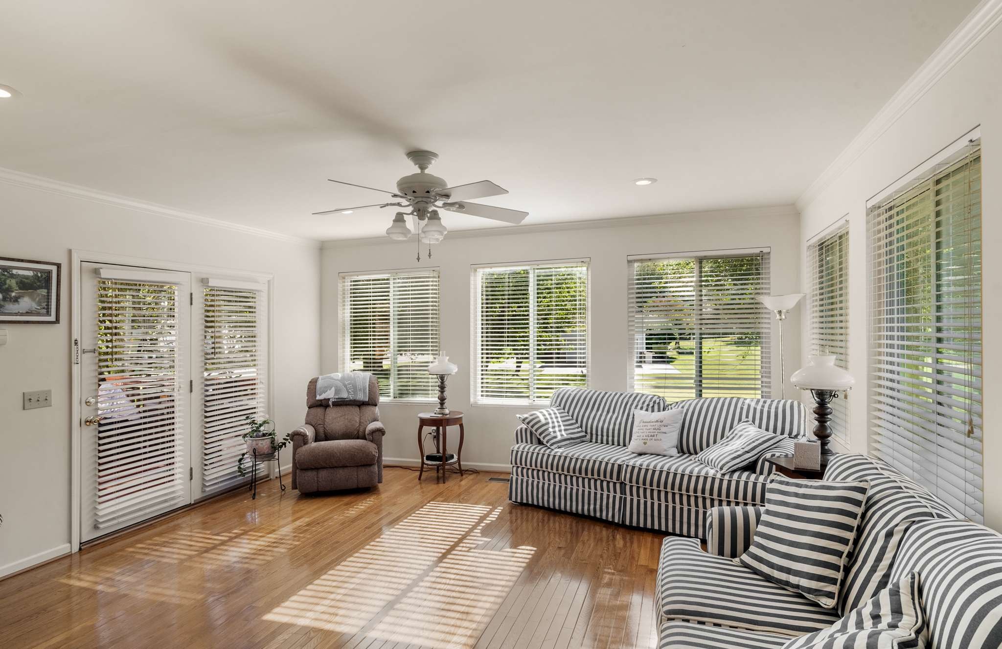 Living room with striped sofas, hardwood floors, and bright windows overlooking a green yard.