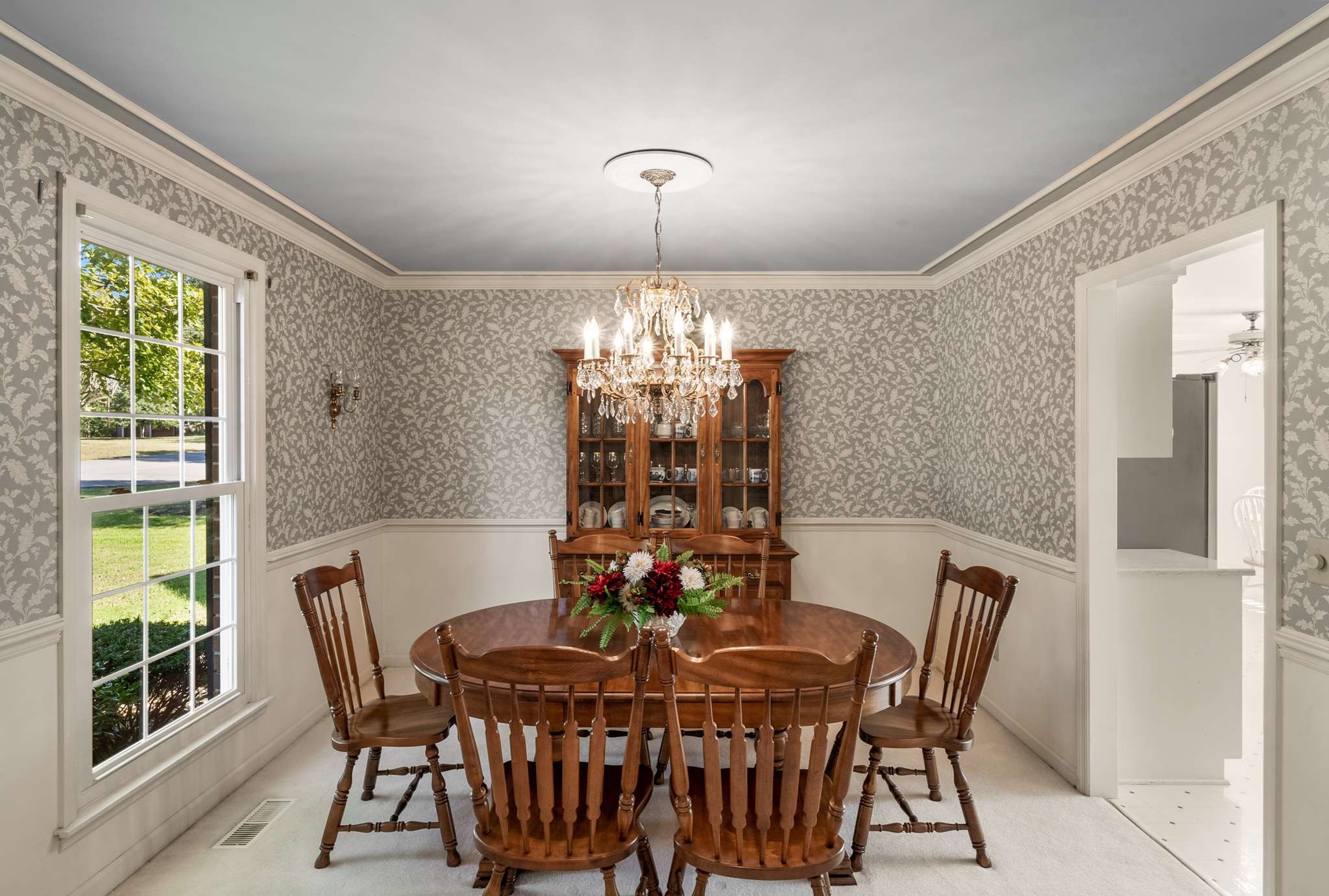 Dining room with a wooden table beneath a crystal chandelier and gray floral wallpaper.