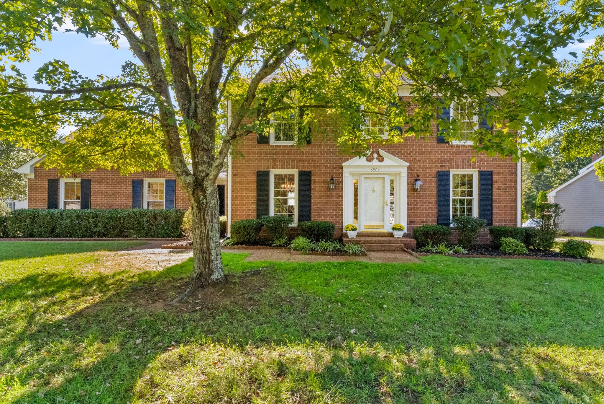 Red brick house with dark shutters, white entry portico, and a large shade tree in the front lawn.