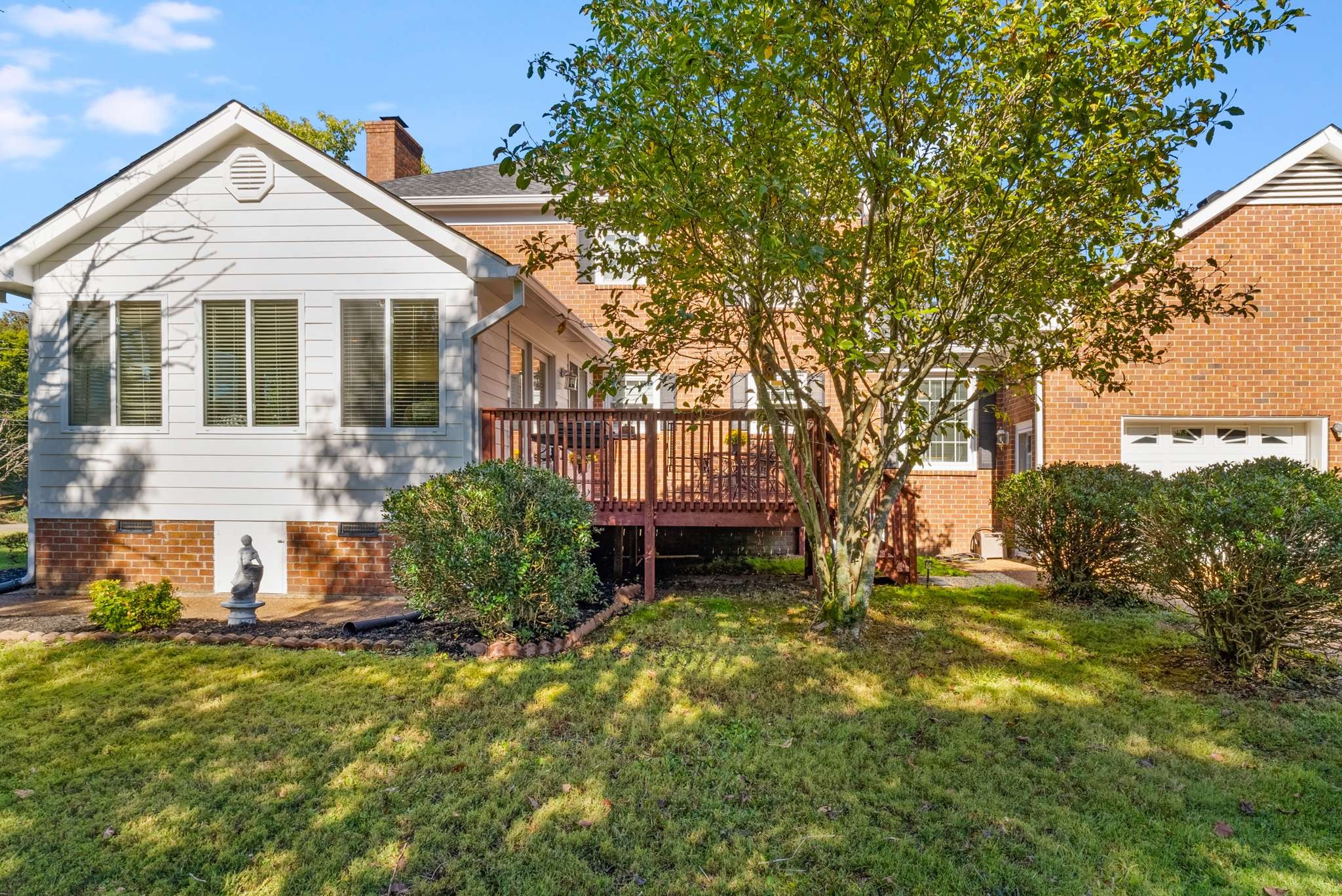 Rear of a brick house with a raised deck, mature tree, and landscaped lawn.
