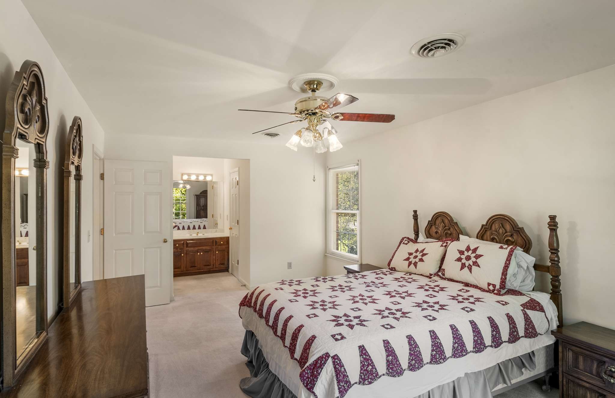 Bedroom with a burgundy-patterned quilt, carved wooden bed, ceiling fan, and adjoining bathroom vanity.
