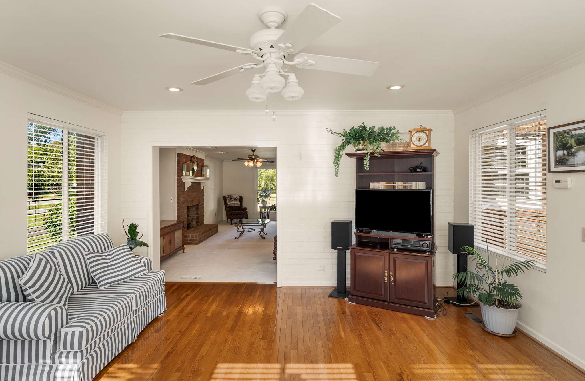 Living room with striped sofa, hardwood floors, and a wide opening to an adjoining room with a brick fireplace.