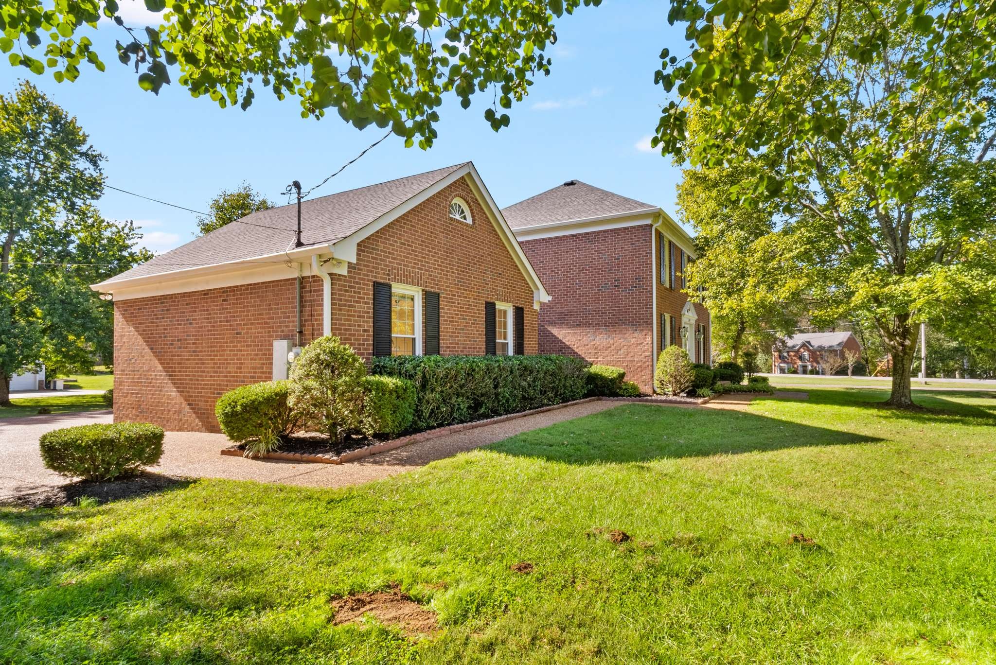 Two-story brick home with black shutters, landscaped shrubs, and a large shaded lawn.