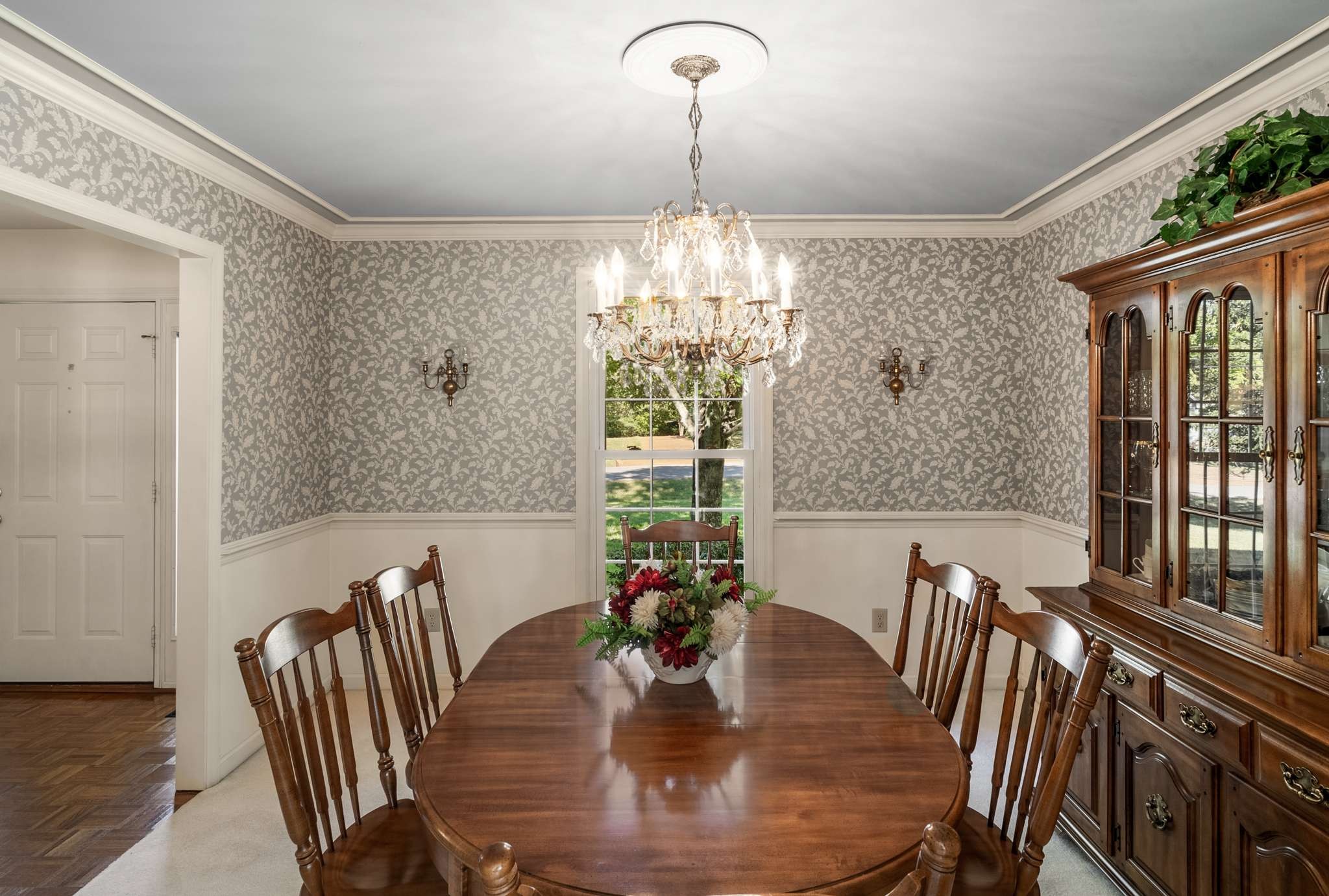 Dining room with a polished oval wood table, crystal chandelier, patterned wallpaper, and glass-front china cabinet.