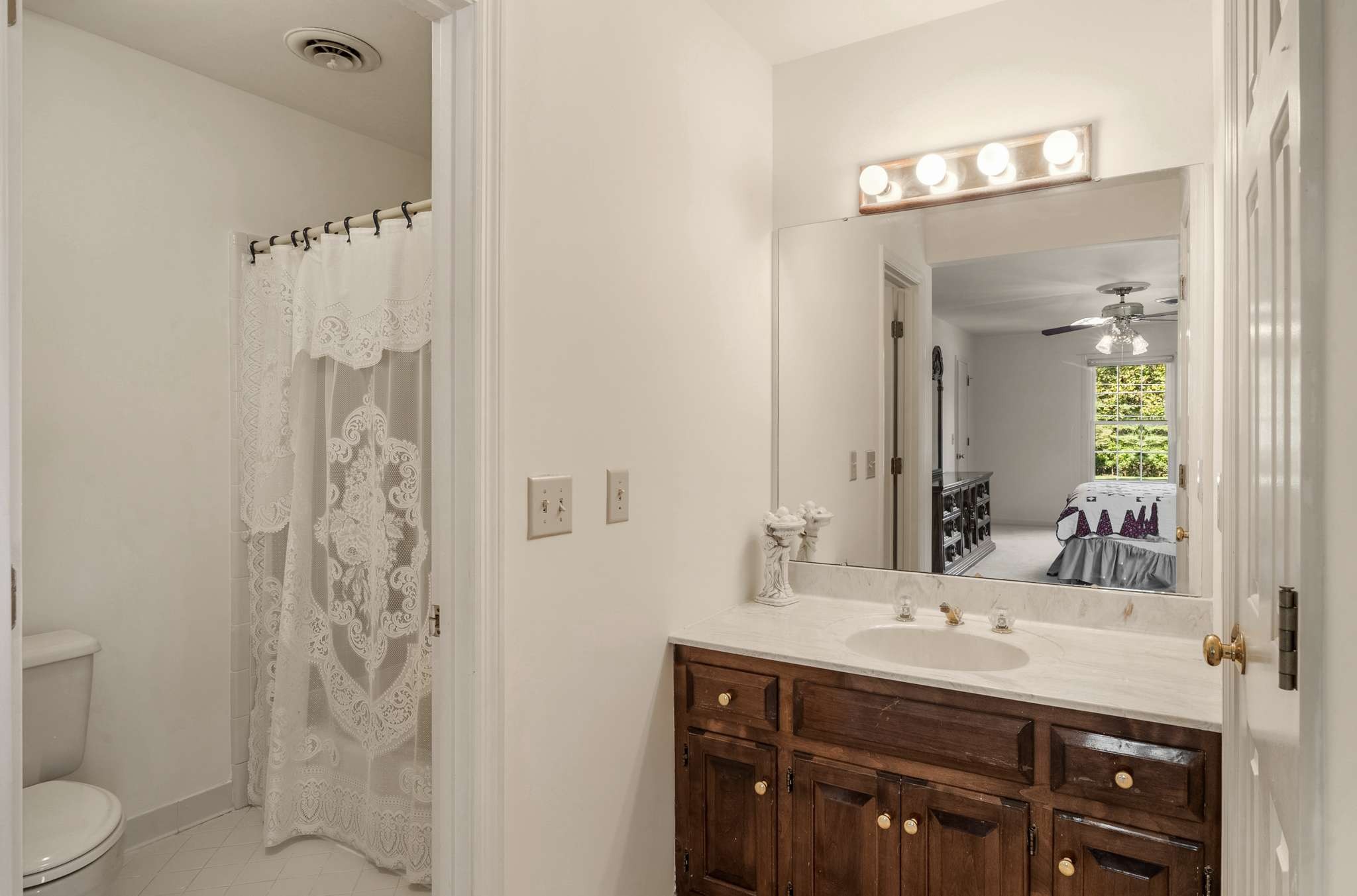 Bathroom with dark wood vanity, wide mirror, lace shower curtain, and toilet; bedroom reflected in the mirror.