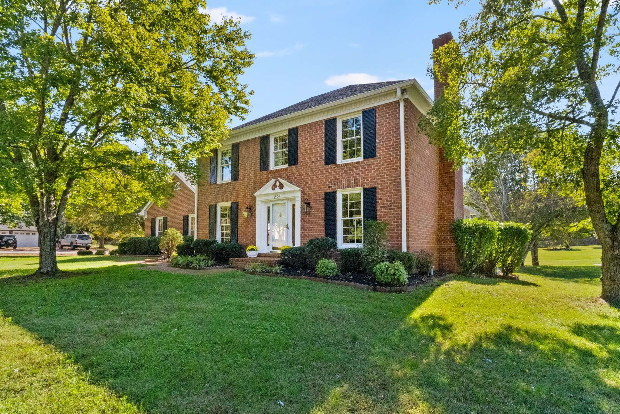 Two-story red-brick house with black shutters, a landscaped entry, and a broad lawn beneath mature trees.