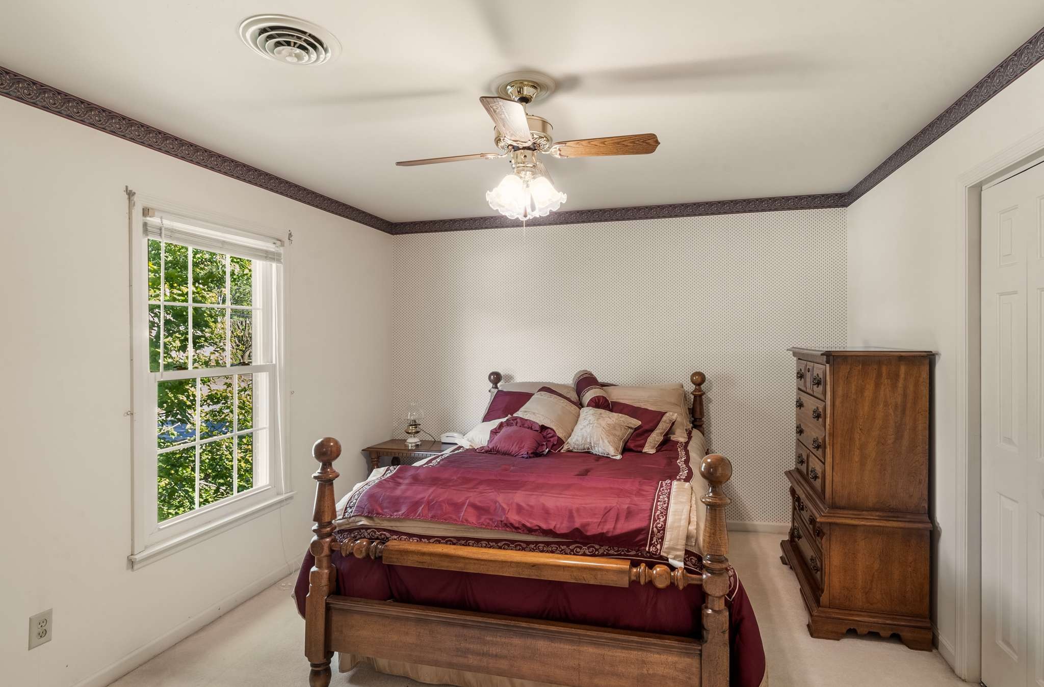 Bedroom with a burgundy-dressed four-poster bed, patterned wallpaper, and a window overlooking leafy trees.