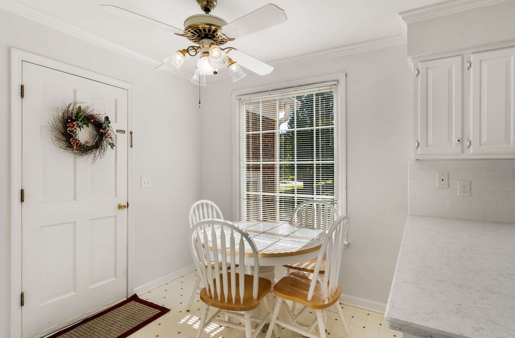 Kitchen breakfast nook with a round white table and chairs beside a sunlit window with blinds.