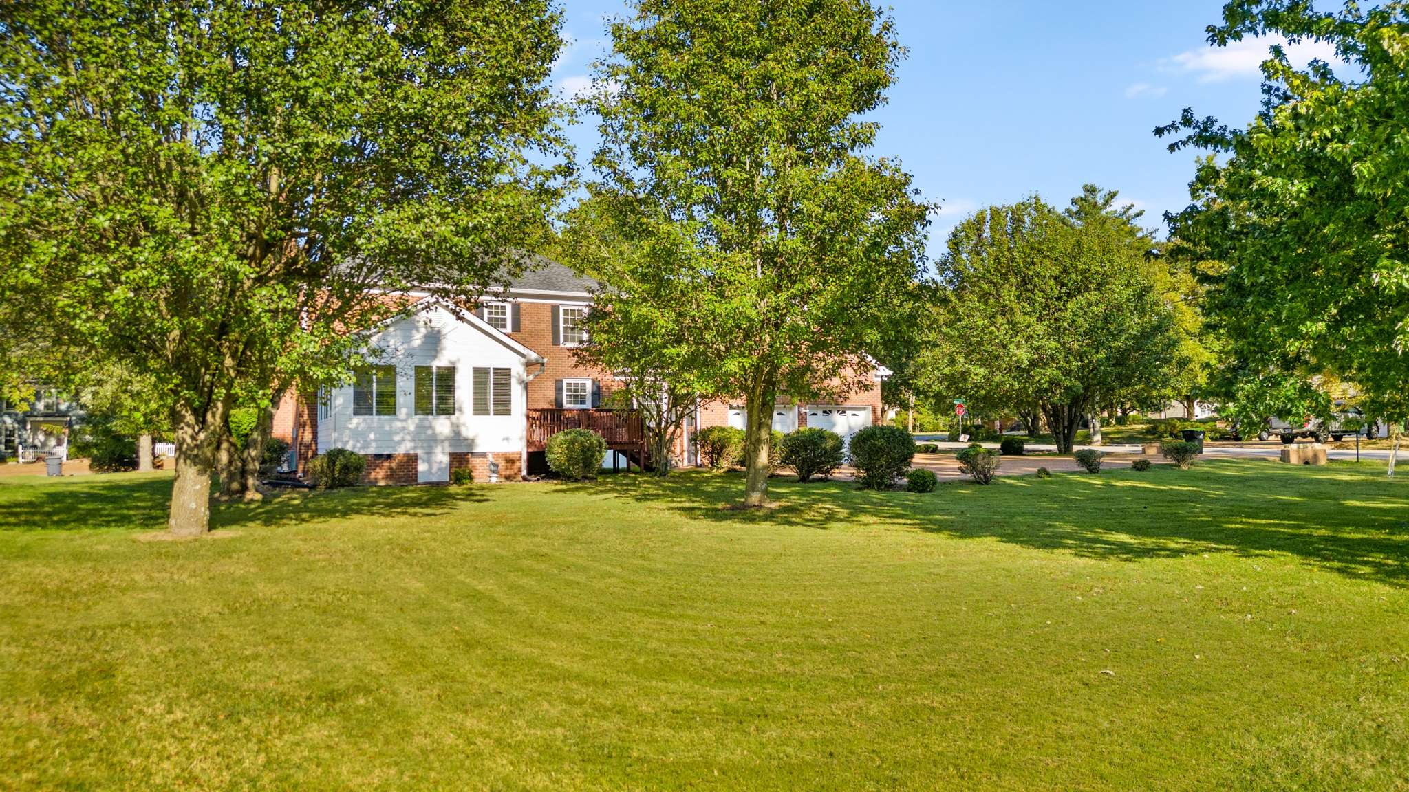 Two-story brick house behind mature trees, with a broad lawn, small deck, and attached garage.