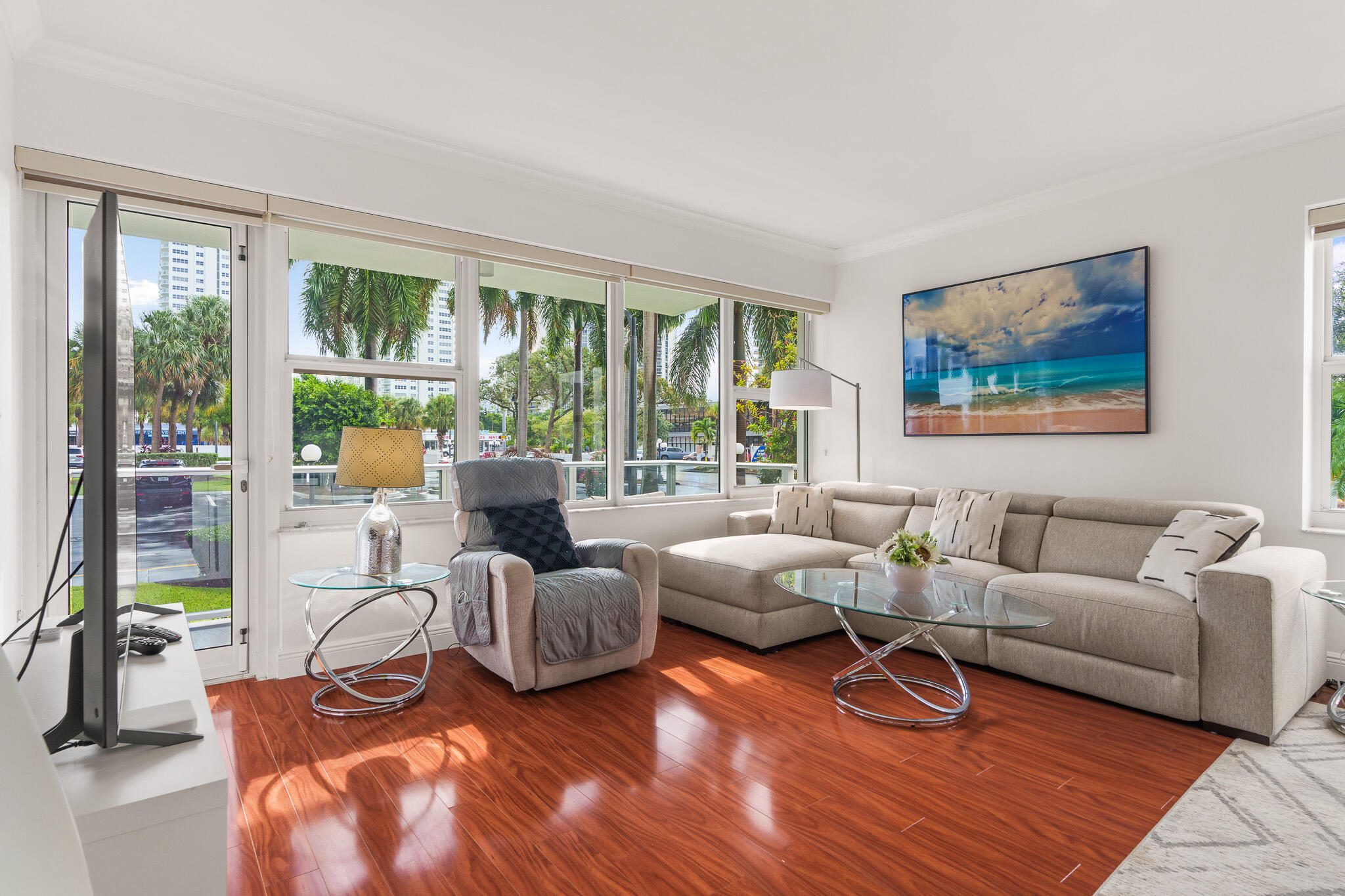 Living room with a beige sectional, glass coffee table, and large windows overlooking palm trees.
