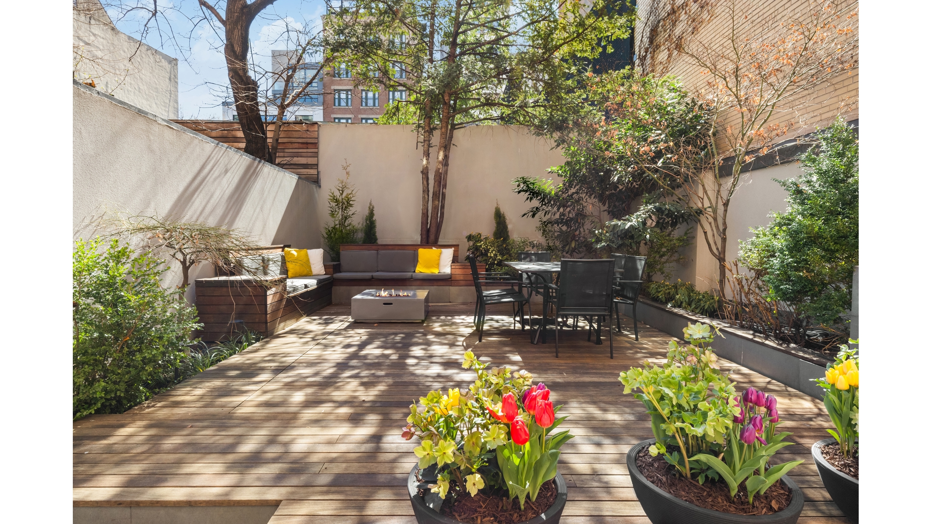 Private terrace with a wooden deck, built-in seating, fire table, dining set, and colorful potted flowers.