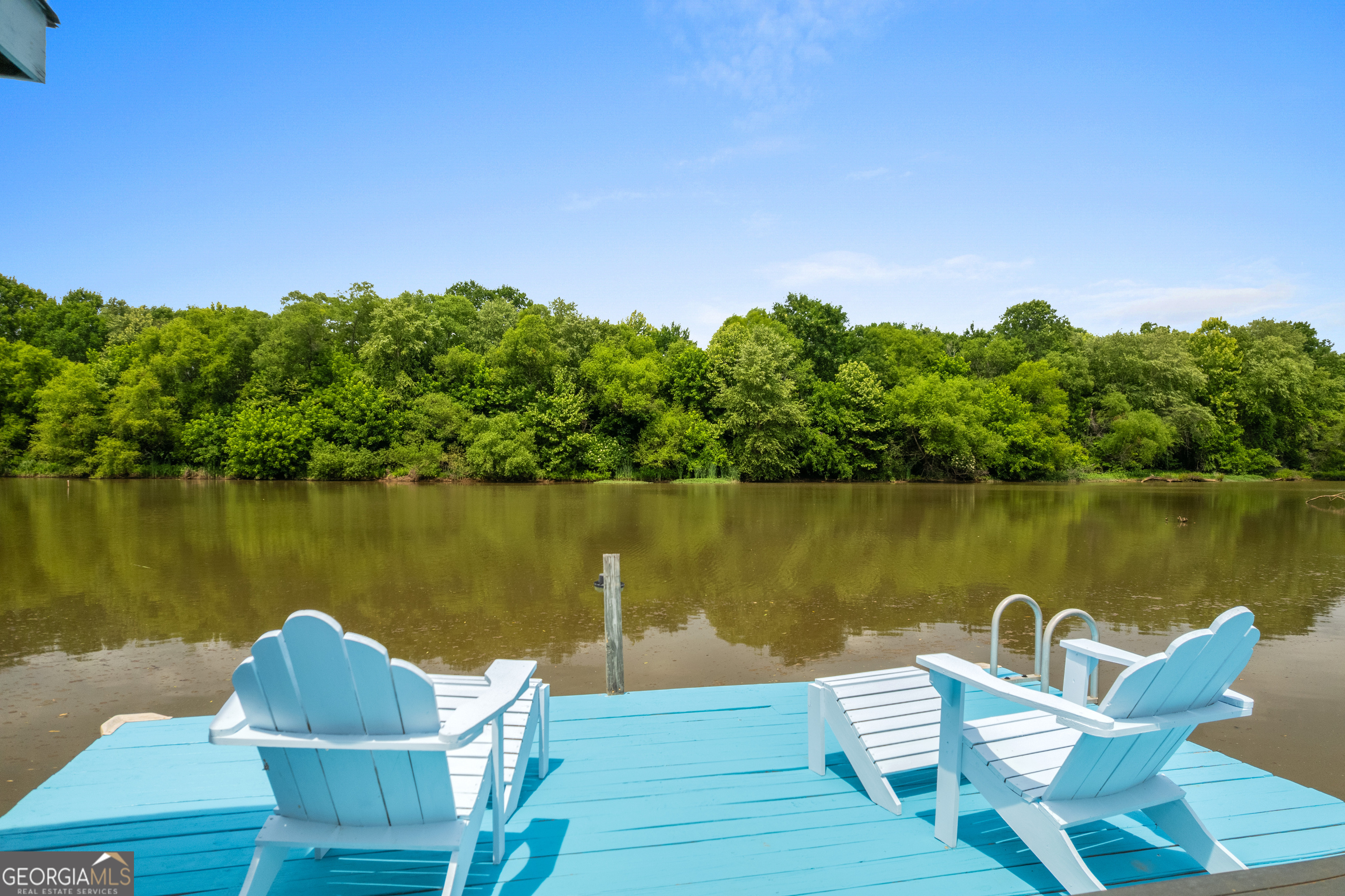 Blue lakeside dock with white Adirondack chairs facing calm water and a wooded shore.