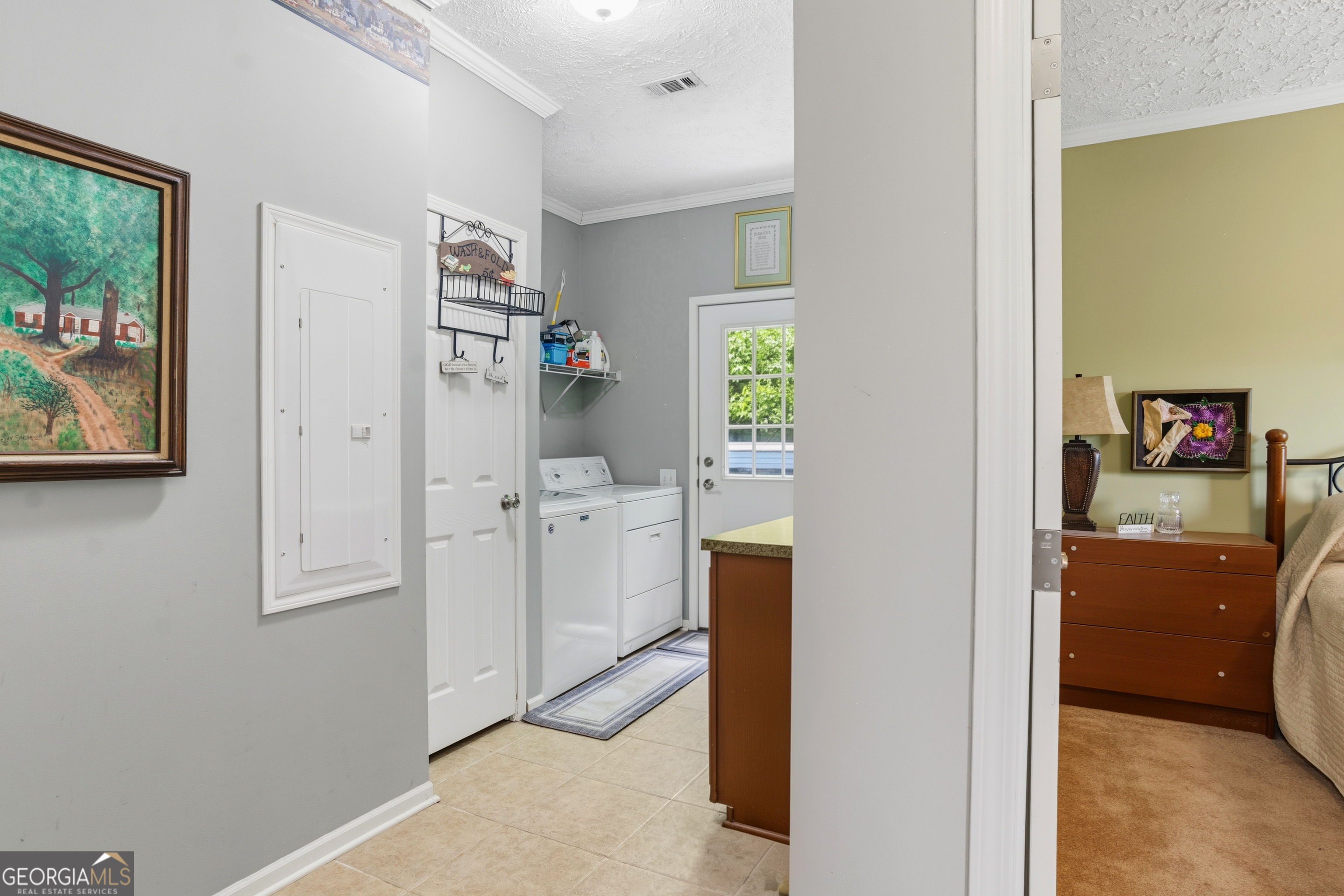 Laundry room with white washer and dryer, wall storage, and tile floor beside a bedroom.