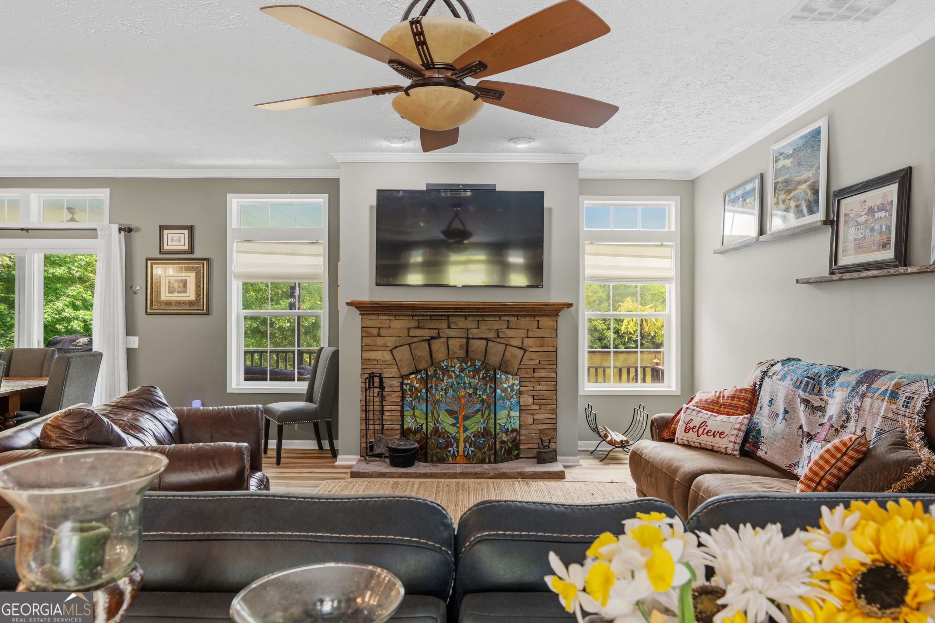 Living room with a stone fireplace, colorful stained-glass screen, mounted television, and ceiling fan.