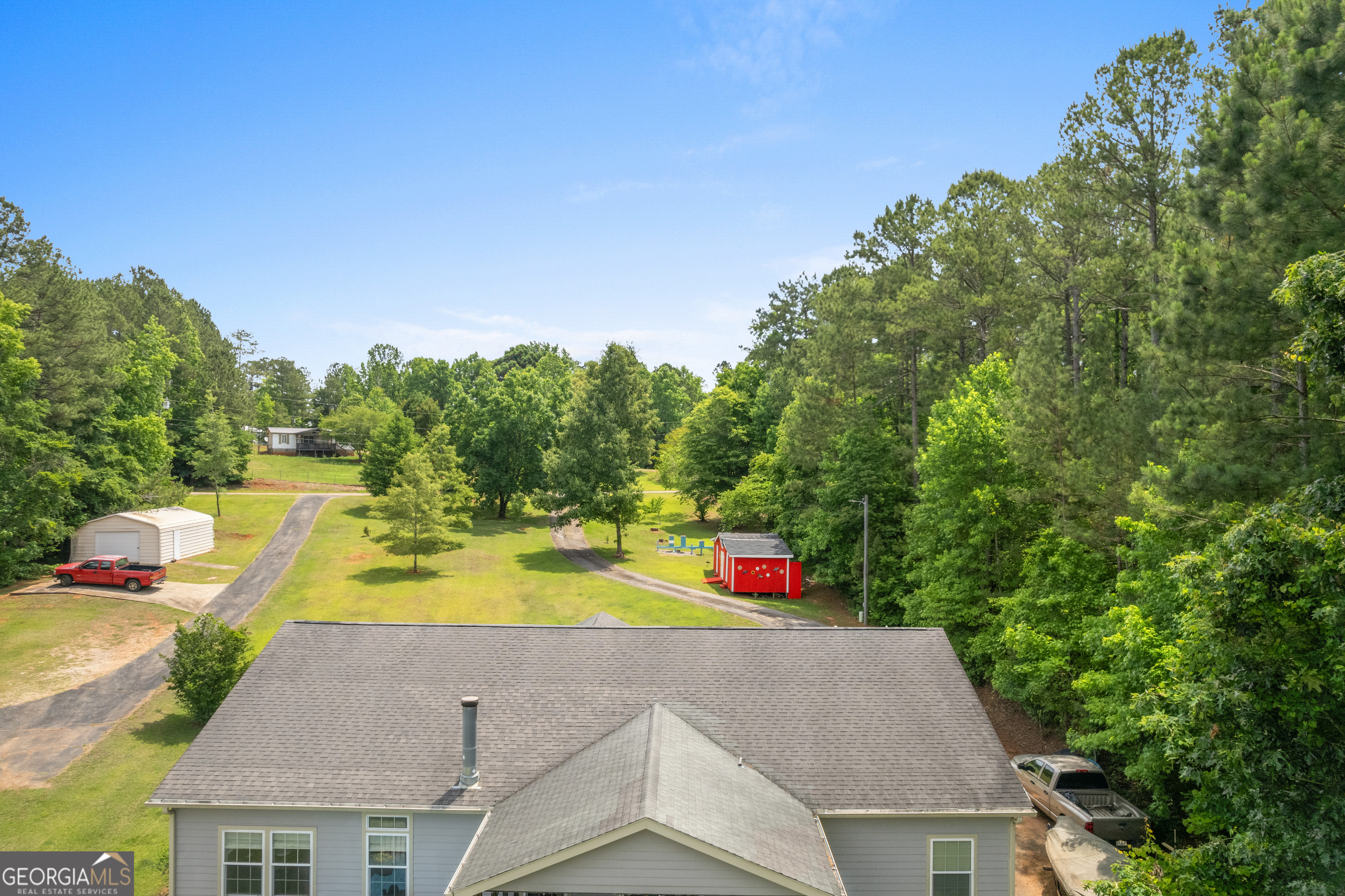 Aerial view of a gray home and wooded grounds with curved driveways, outbuildings, and parked vehicles.