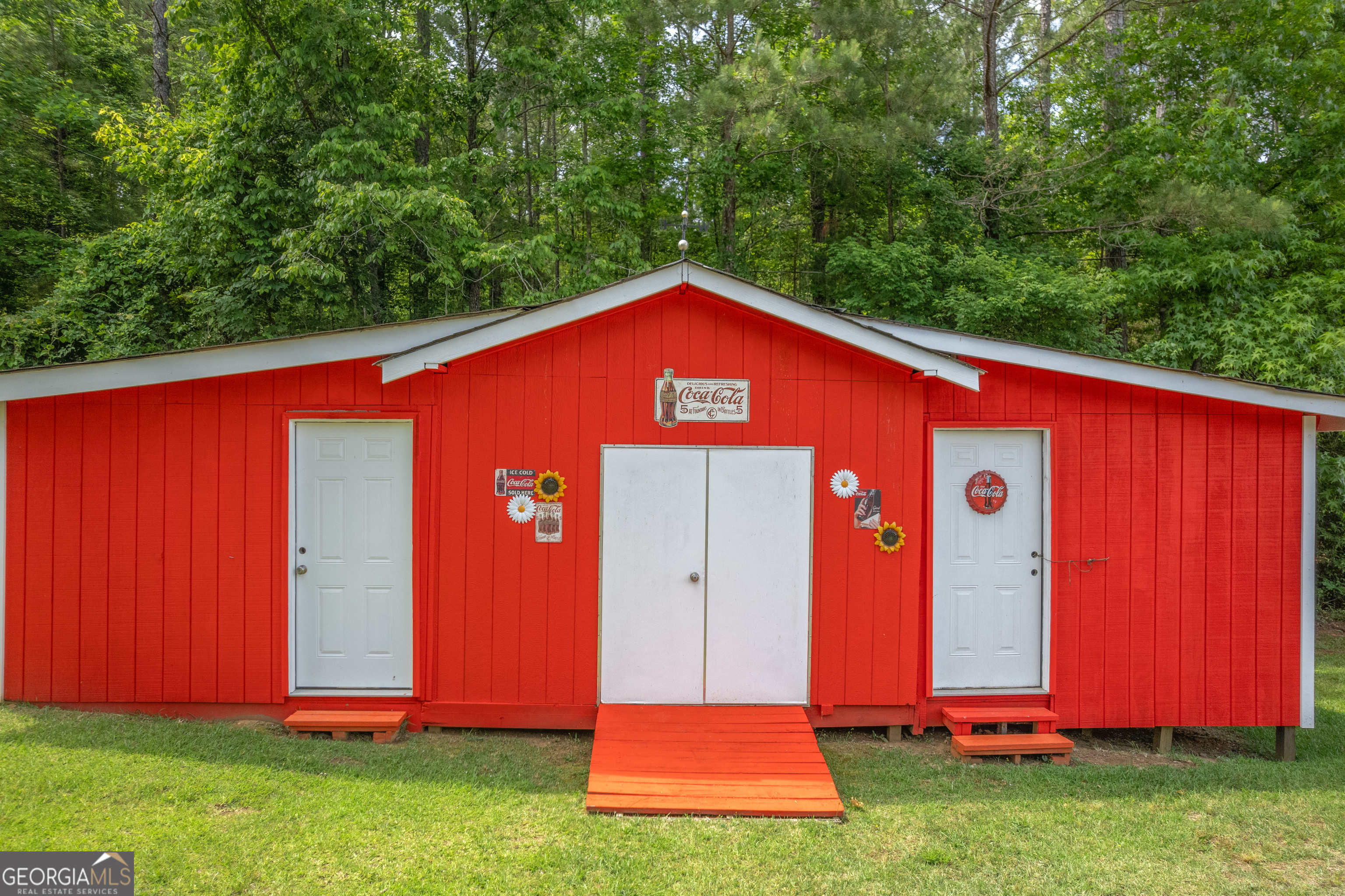 Red outbuilding with three white doors and a ramp, set against dense trees.