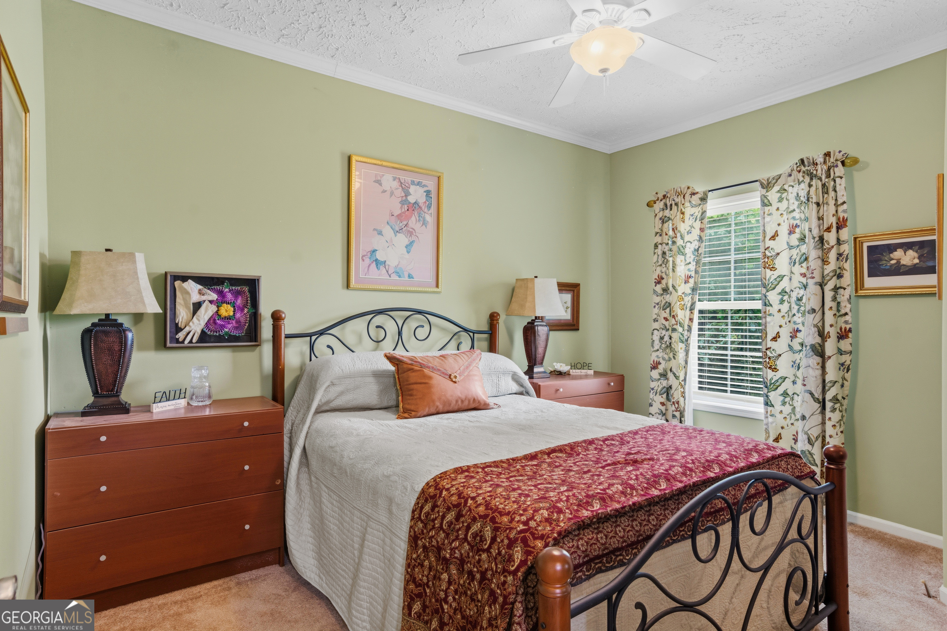 Bedroom with a wrought-iron bed, red patterned bedspread, and floral curtains framing a shuttered window.