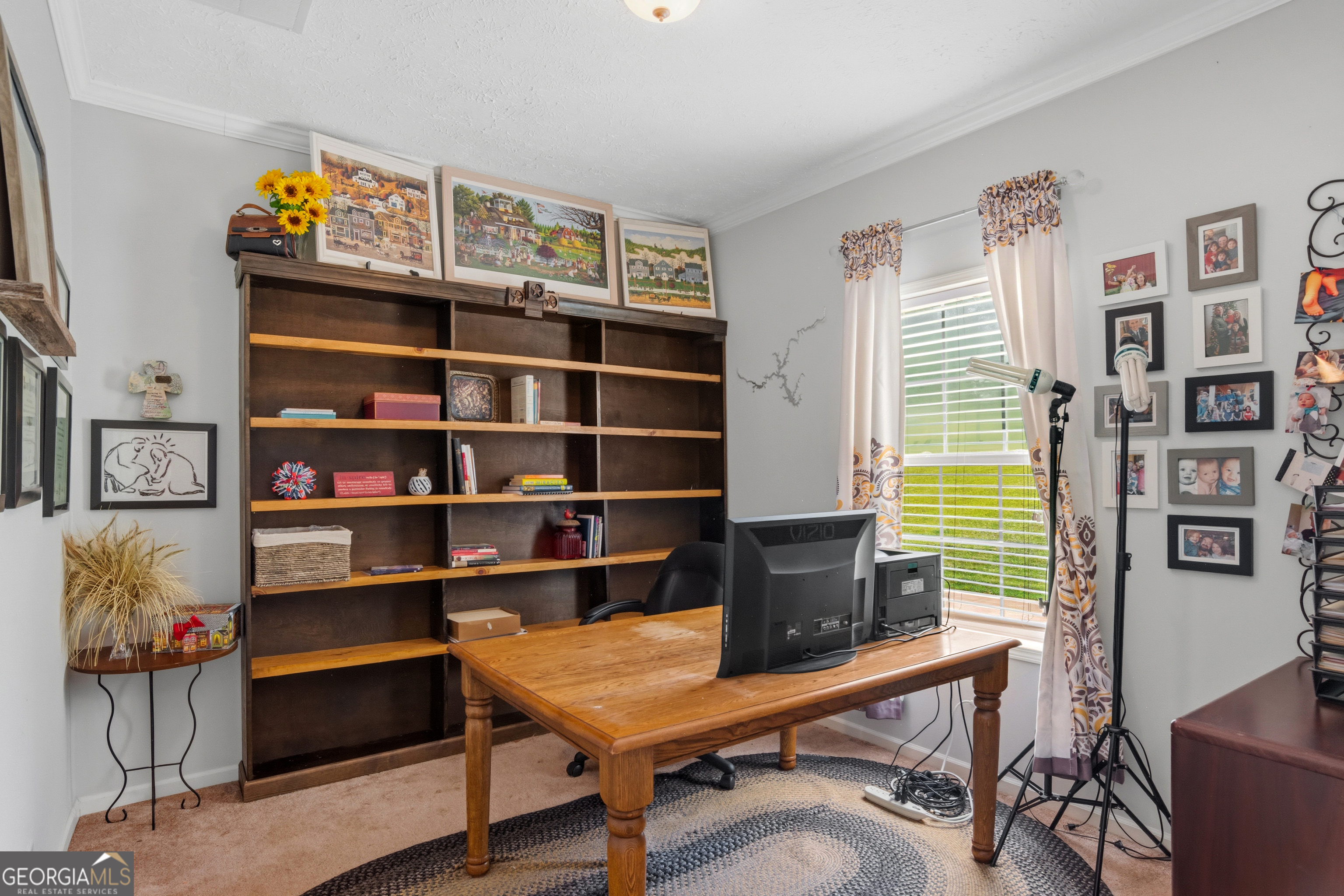Furnished home office with a wooden desk, large bookcase, and bright curtained window.