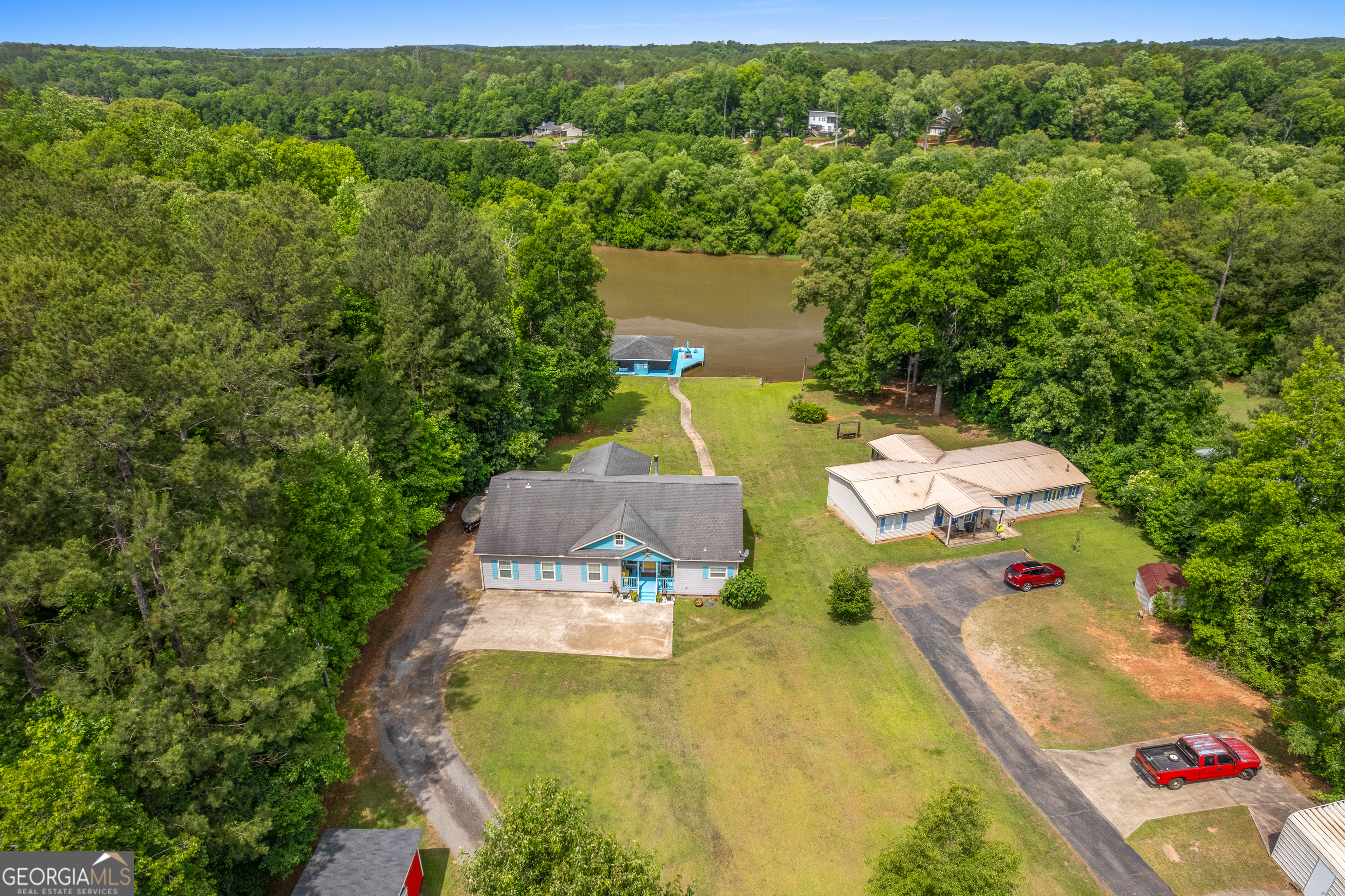 Aerial view of two houses beside wooded waterfront, with a walkway leading to a blue waterside structure.