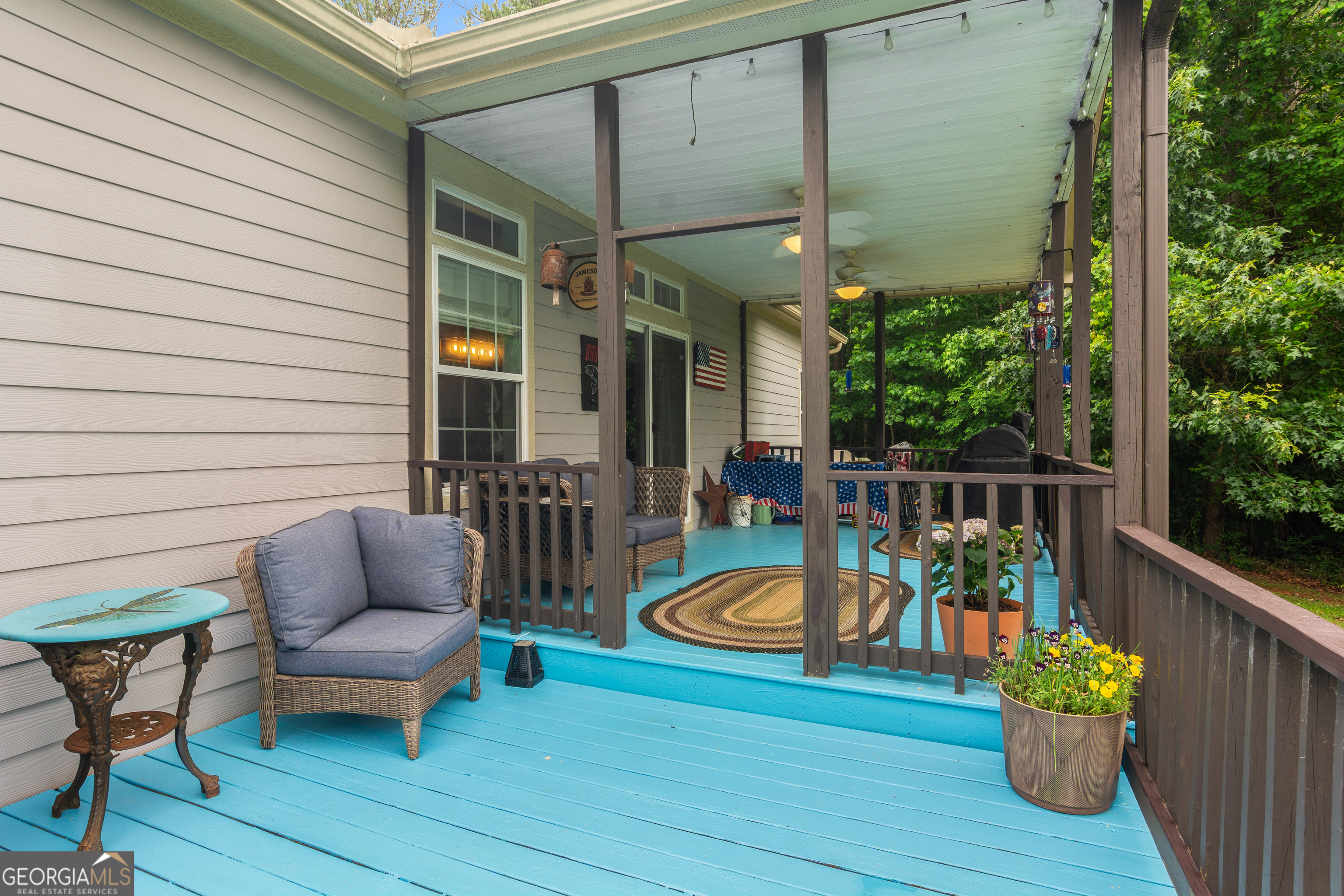 Covered porch with turquoise decking, wicker seating, and potted flowers among leafy trees.