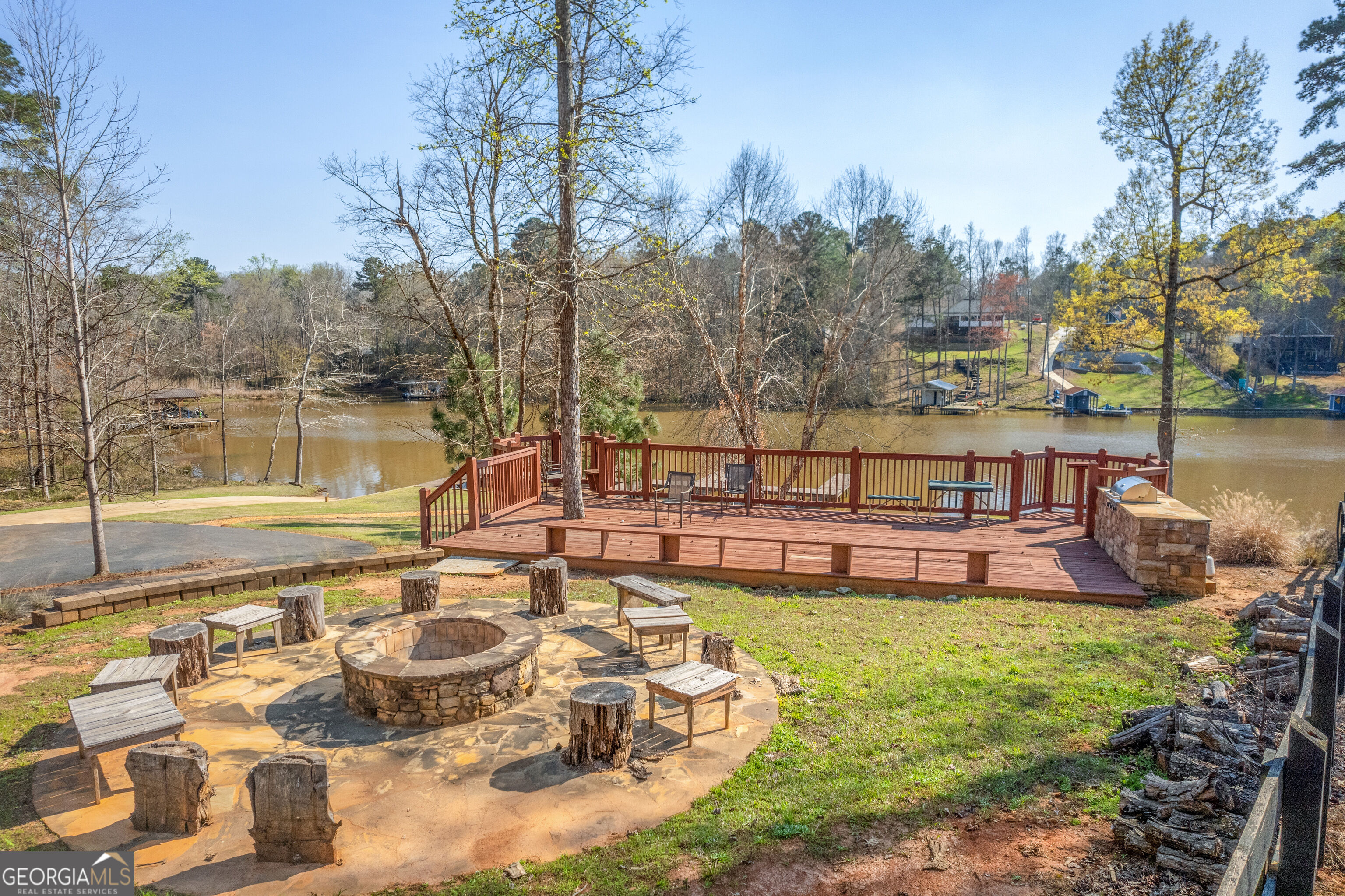 Waterfront deck and stone fire pit patio overlooking the water, with seating and trees along the shore.