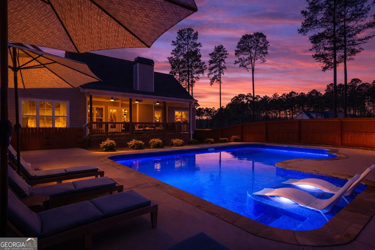 Backyard pool and patio with lounge chairs and umbrellas at dusk, beside an illuminated house.