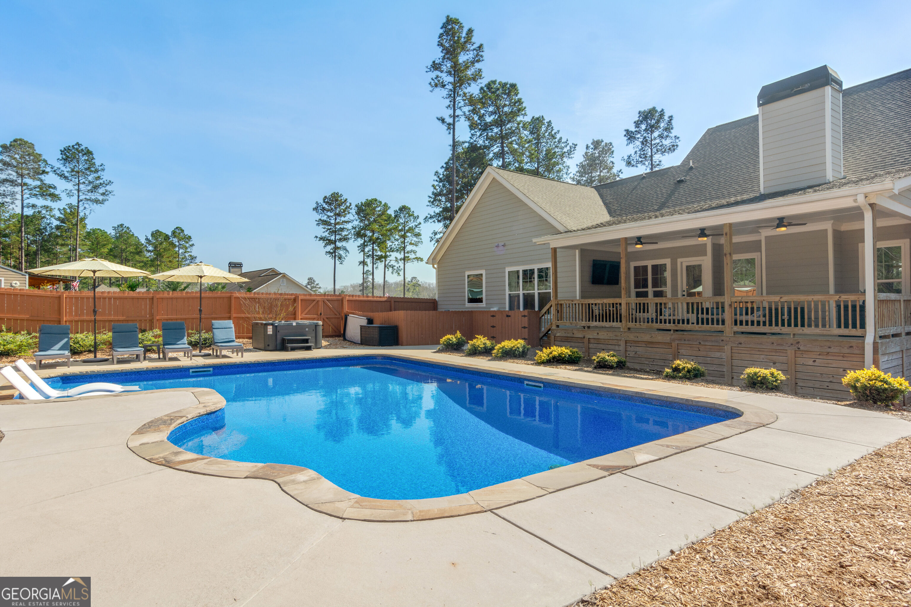 Backyard swimming pool beside a gray house, with a covered porch, lounge chairs, umbrellas, and hot tub.