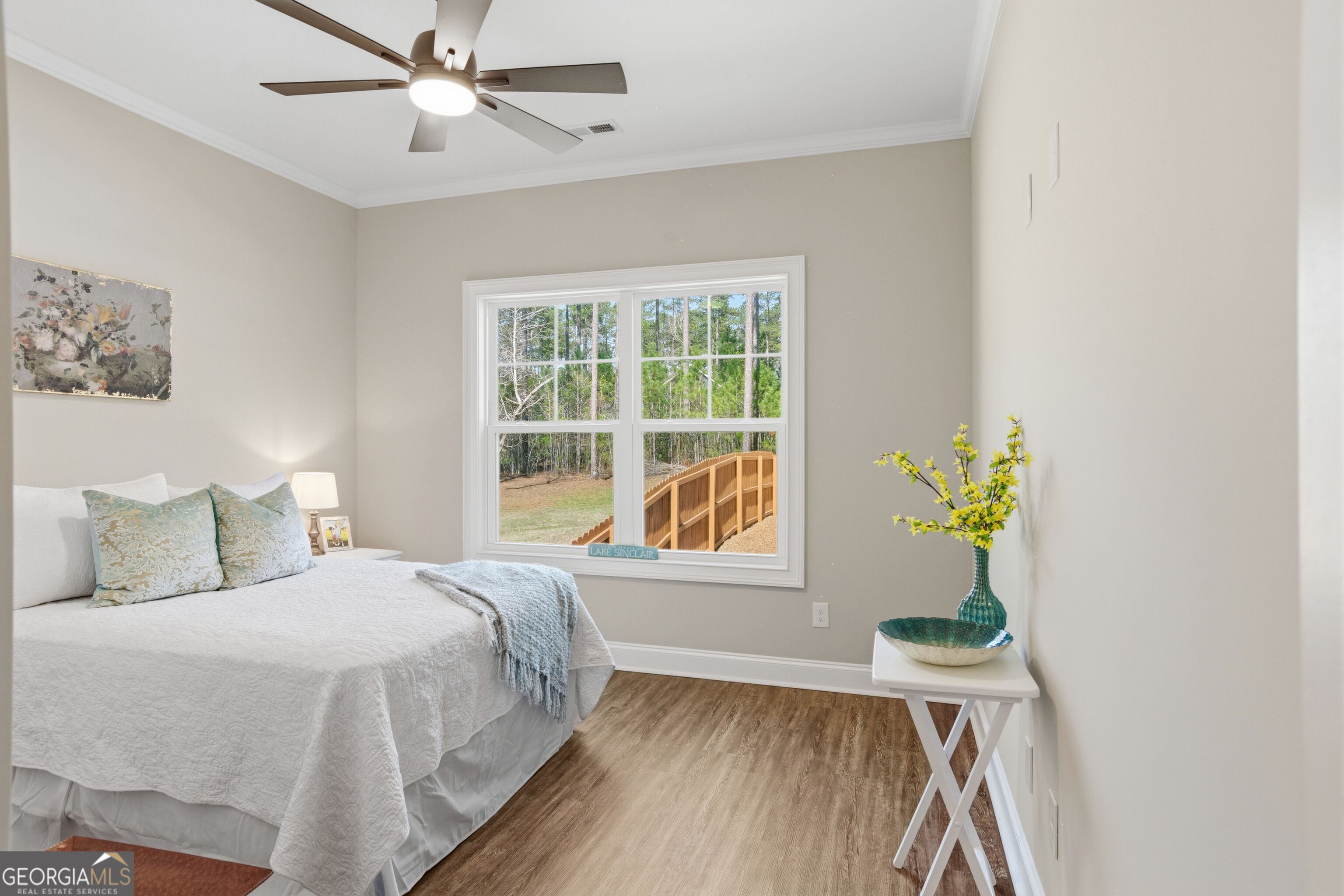Staged bedroom with a white bed and a large window overlooking trees and a wooden fence.