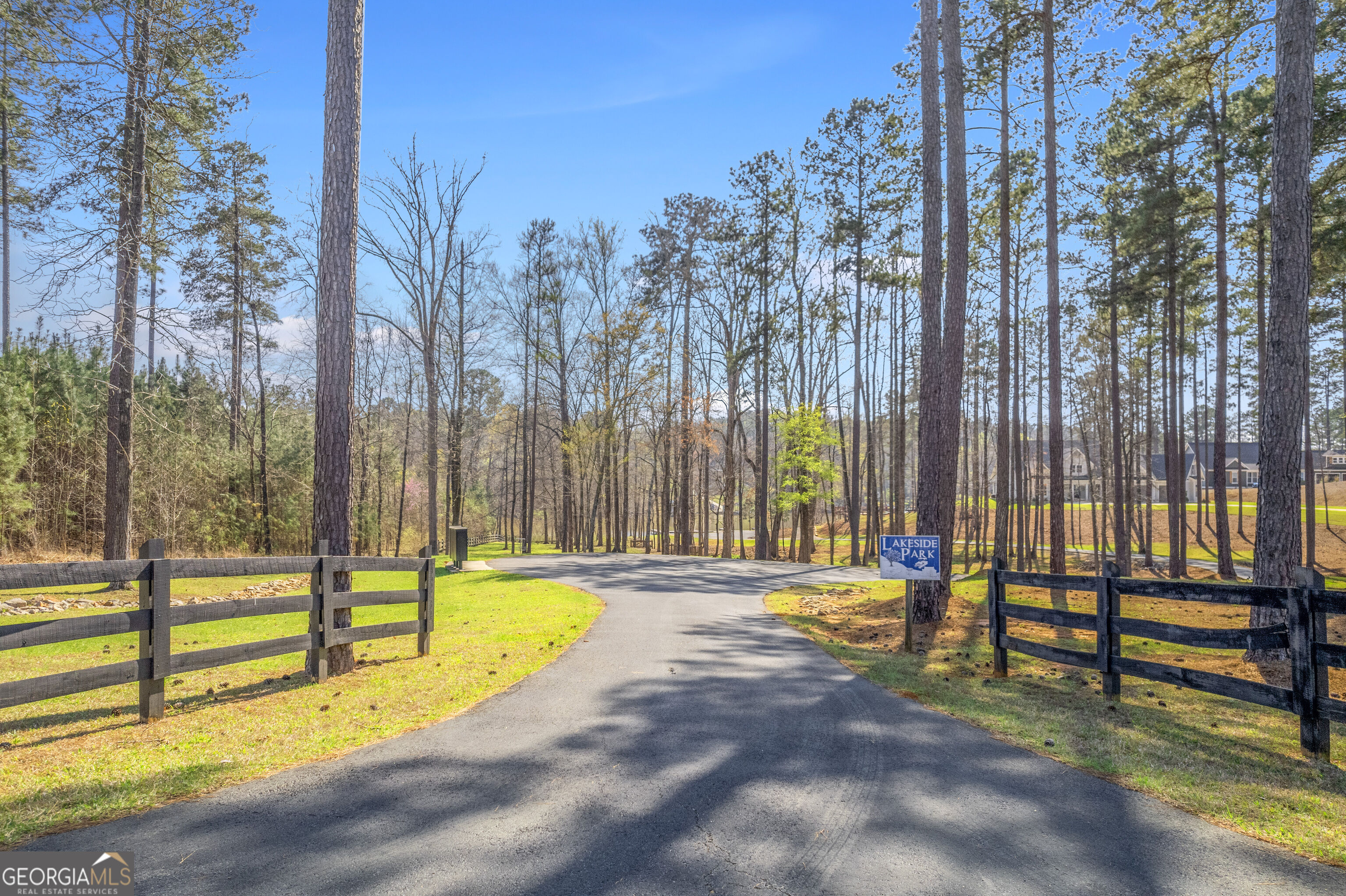 Wooded Lakeside Park entrance with a curving road, dark fences, and a roadside sign.