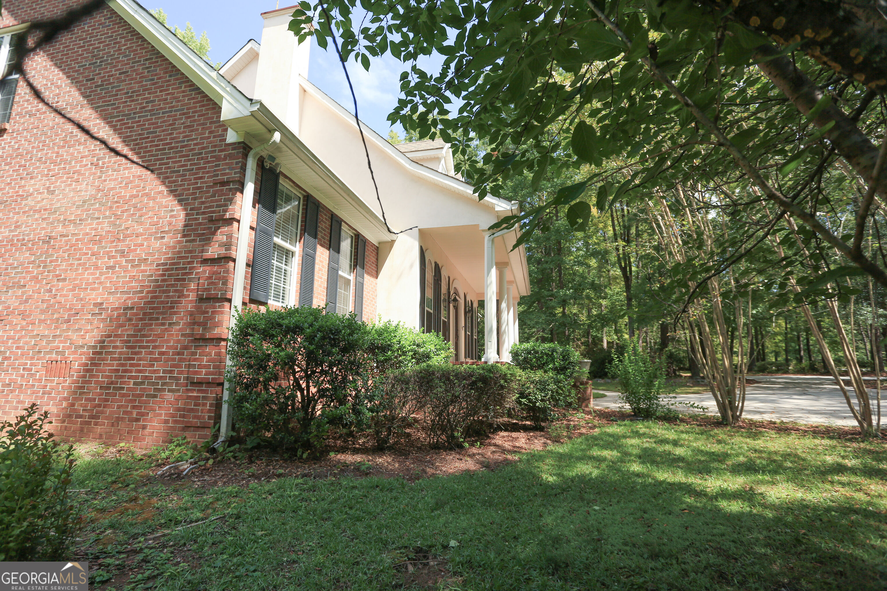 Brick-and-siding home with black shutters, landscaped shrubs, and a wooded driveway.