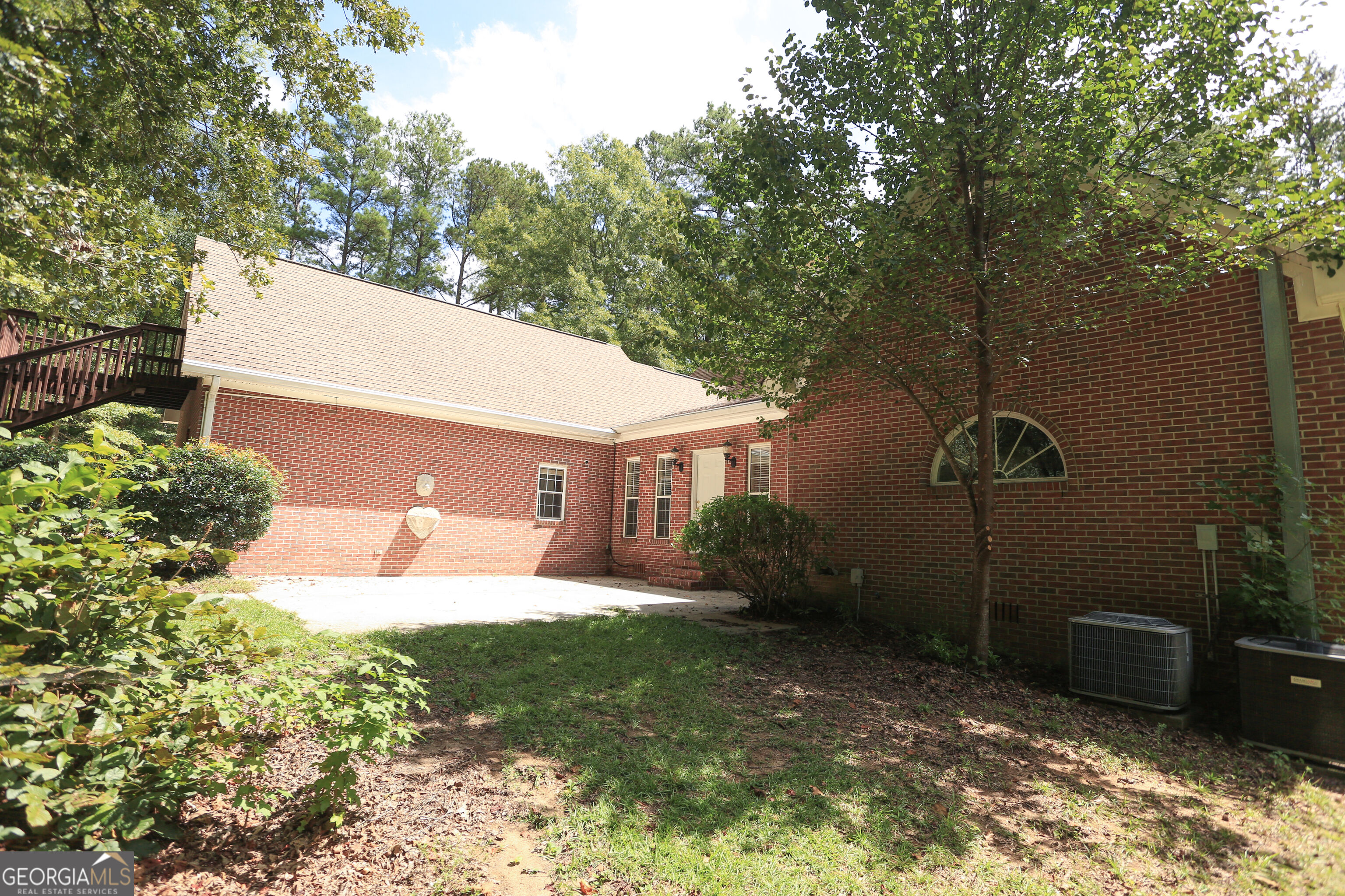 Red-brick house rear with a concrete patio, raised deck stairs, and mature trees.