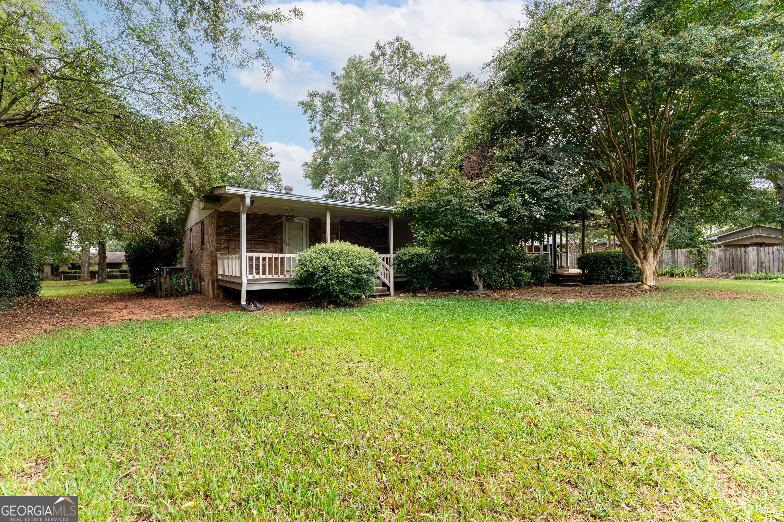 Brick house with a covered porch and white railing beneath mature trees, beyond a broad lawn.
