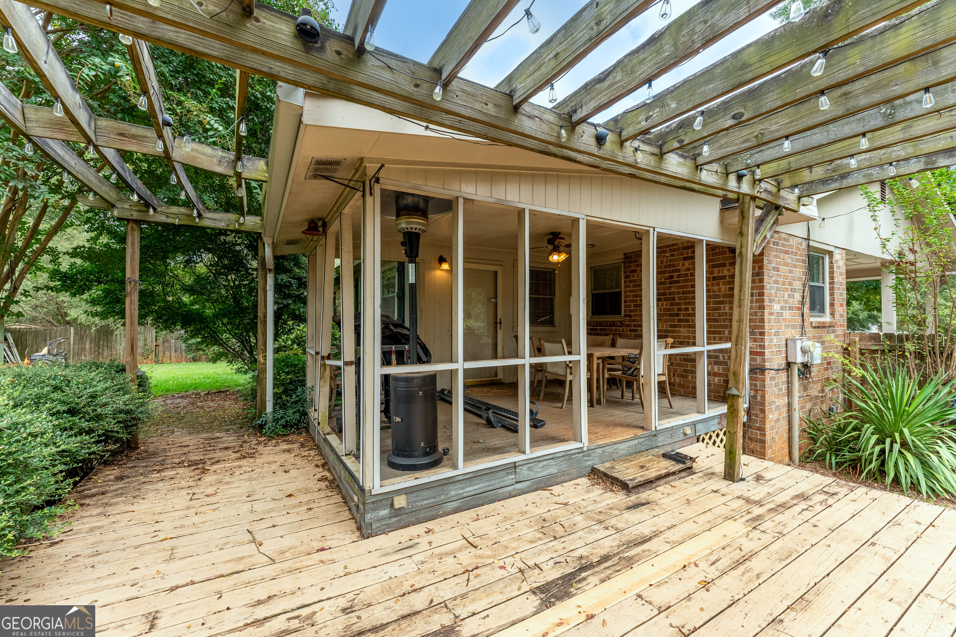 Wooden deck and screened porch beneath a pergola with string lights and surrounding greenery.