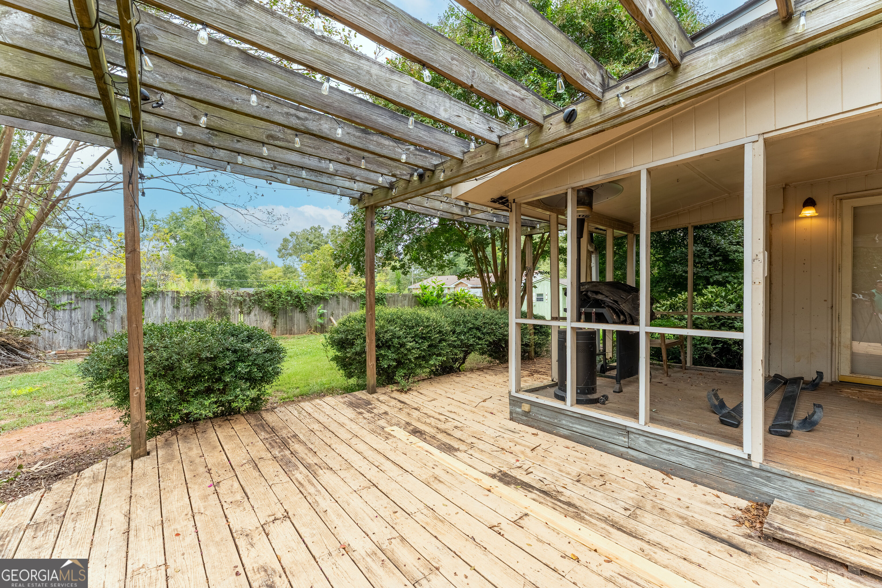 Wooden deck beneath a pergola with string lights, overlooking a fenced backyard beside a screened porch.
