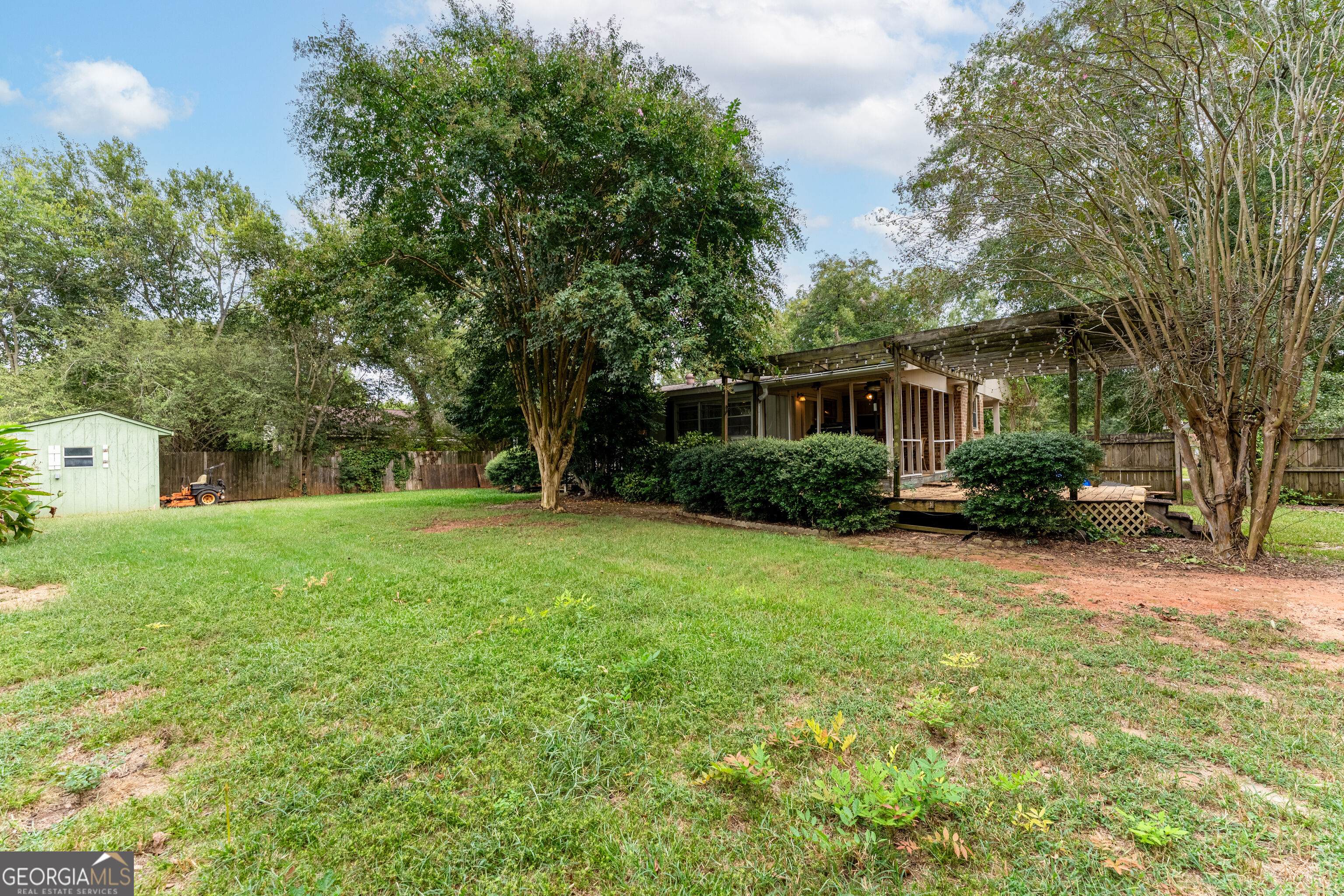 Home’s backyard with a covered deck, pergola, mature trees, lawn, and small shed.