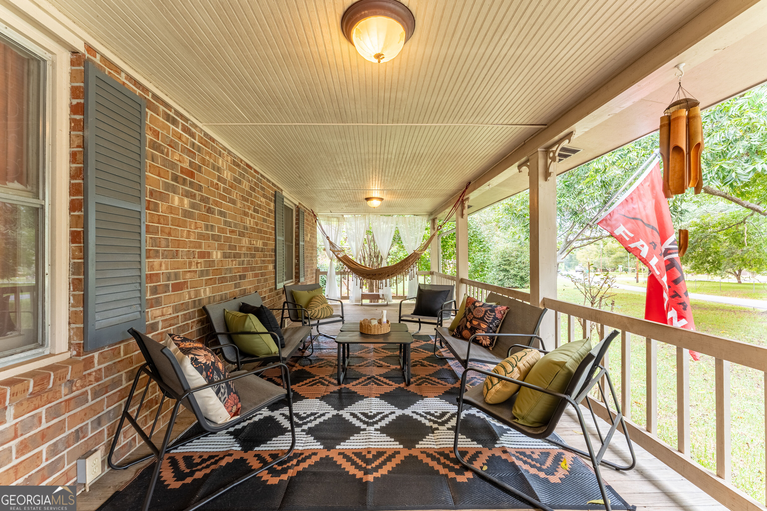 Covered brick porch with cushioned seating, patterned rugs, a hammock, and a red FOR SALE flag outside.